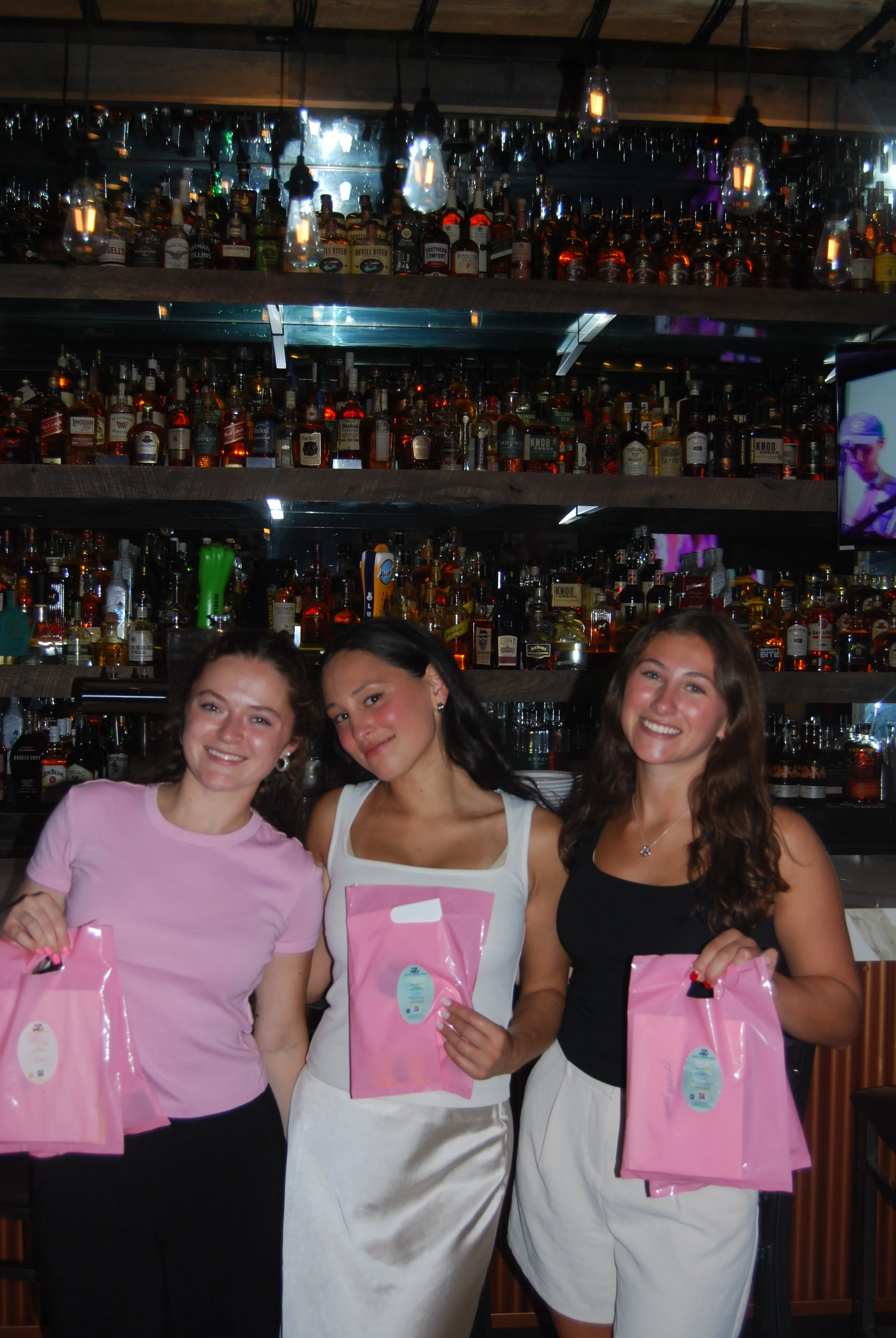 Three women smiling and holding pink gift bags standing in front of a bar with bottles of alcohol on shelves.