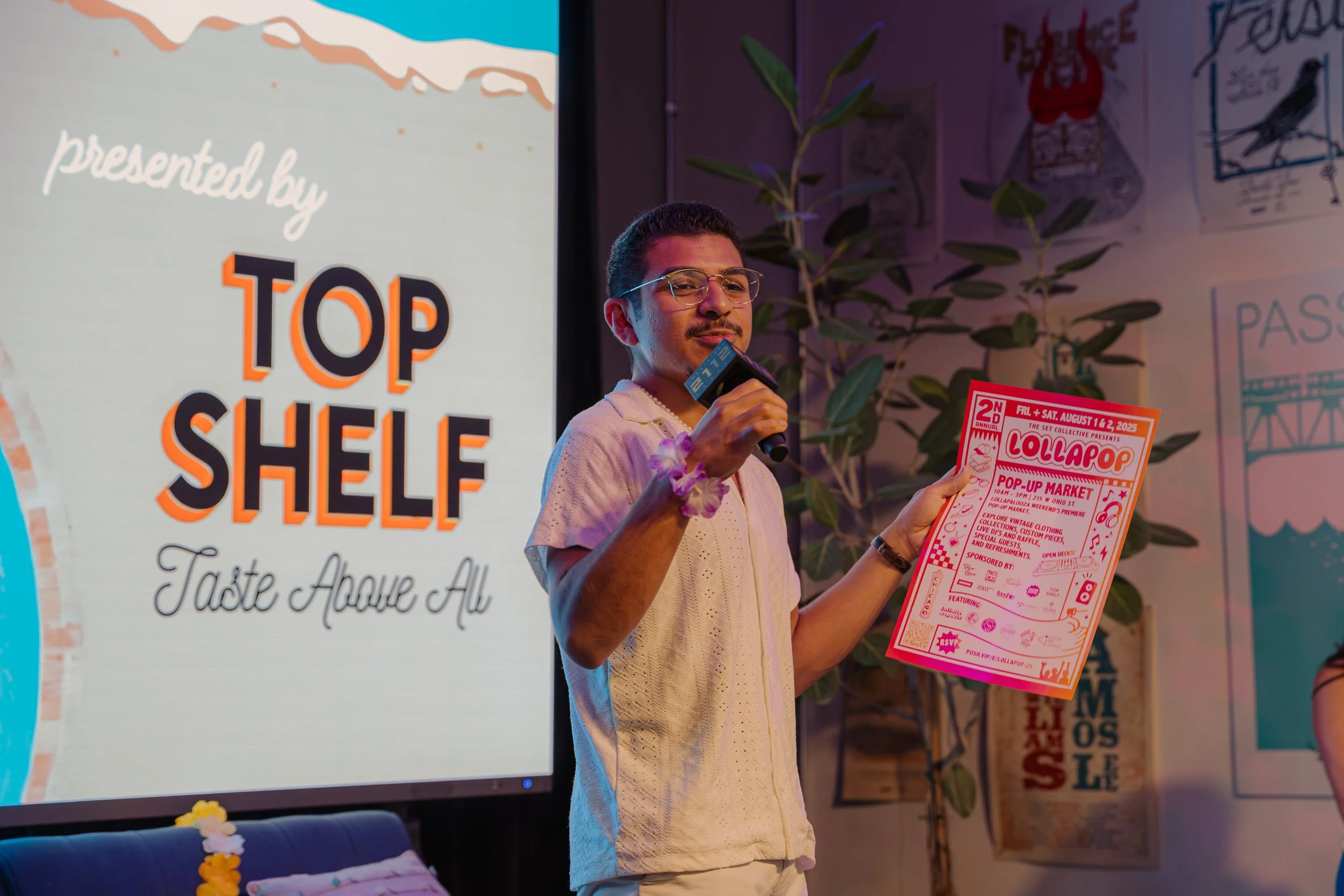 A man with glasses and a white shirt holding a microphone and a pink flyer, standing next to a projection screen that reads "presented by TOP SHELF Taste Above All" in a decorated indoor space with a plant in the background.