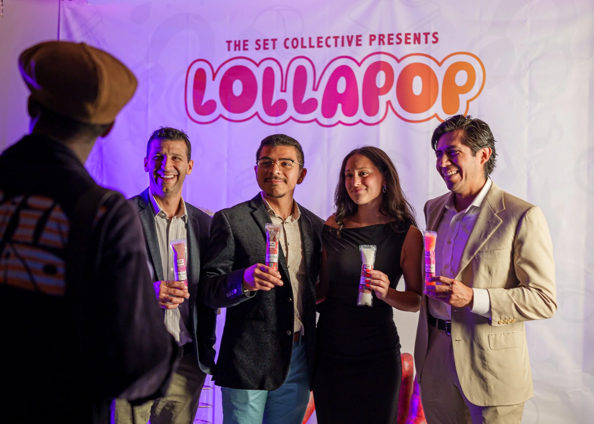 Group of four people at a party holding glow sticks, standing in front of a backdrop with the words 'Lollapalooza' and 'The Set Collective Presents,' smiling and engaging in conversation.
