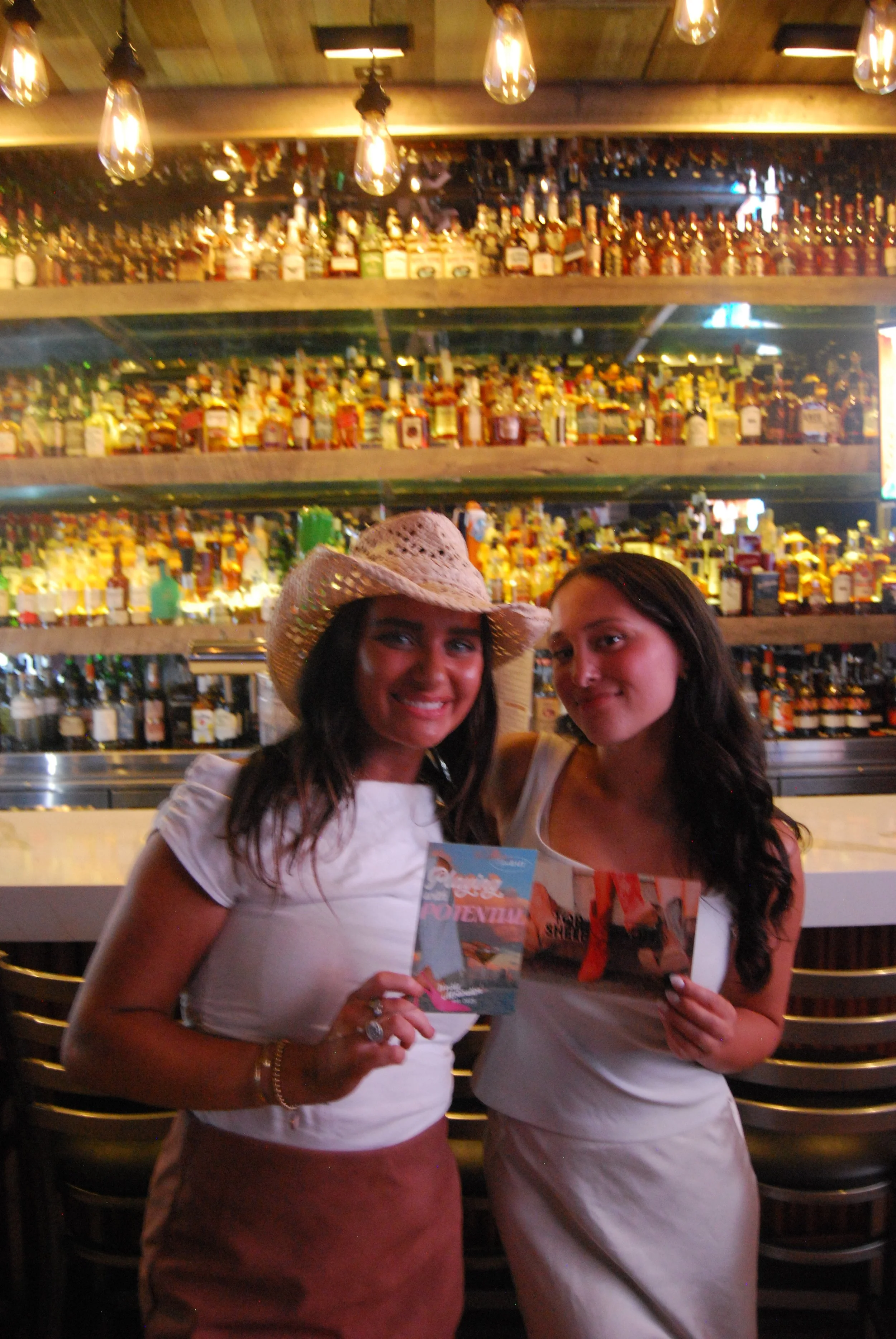 Two women smiling at a bar, holding colorful cards or brochures, with shelves of various bottles of liquor behind them in a dimly lit, stylish setting.