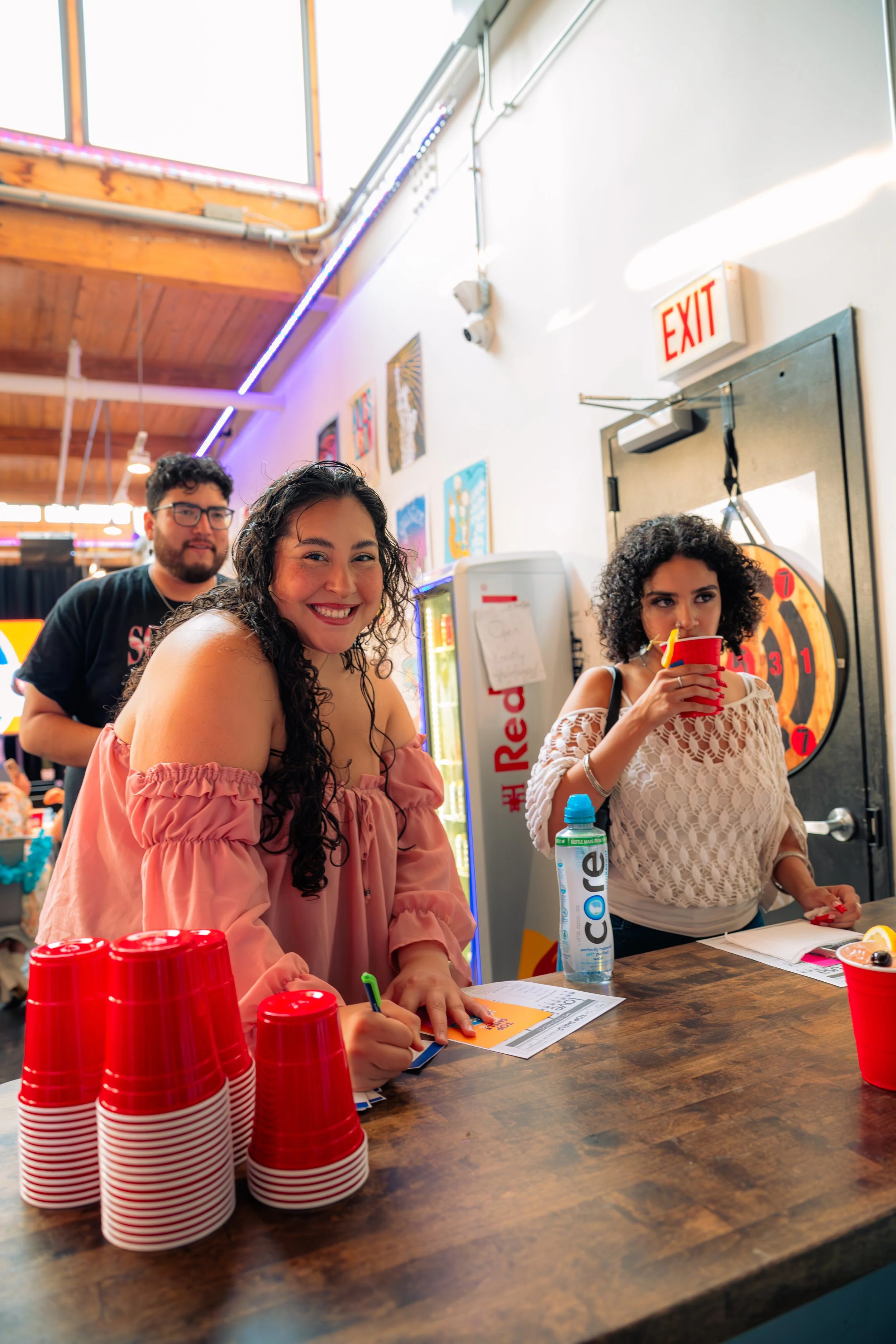 People at a bar signing a sheet of paper, with red cups stacked on the counter and a woman drinking from a red cup, inside a room with wooden ceiling and colorful wall art.