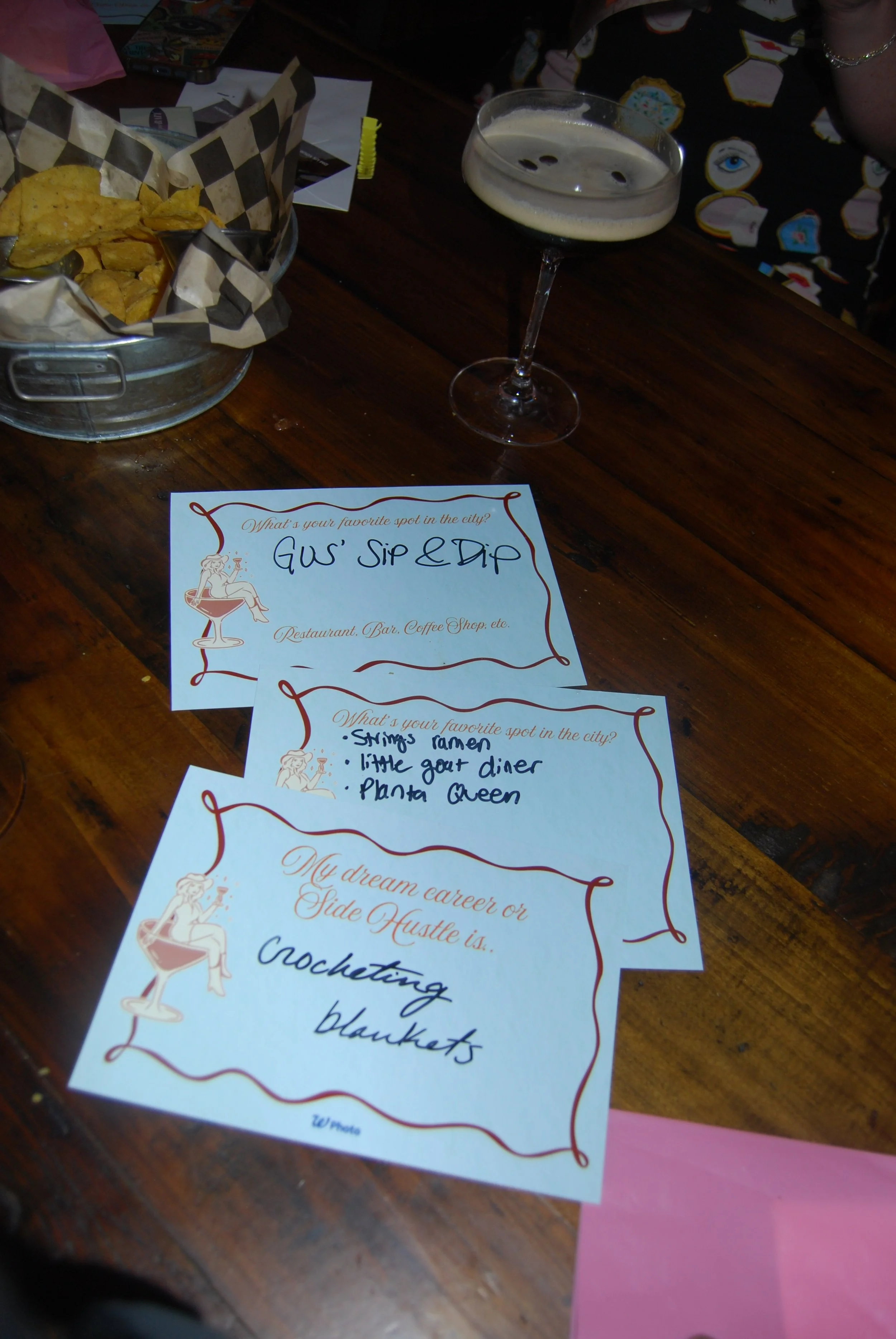 Two handwritten notes on decorative paper, a glass of dark beer, and a basket of tortilla chips on a wooden table. The notes ask about favorite spots in the city, list favorite spots including 'Shops Famer,' 'The Gut Diner,' and 'Planta Queen,' and mention a dream career or side hustle of 'croteting blankets.'