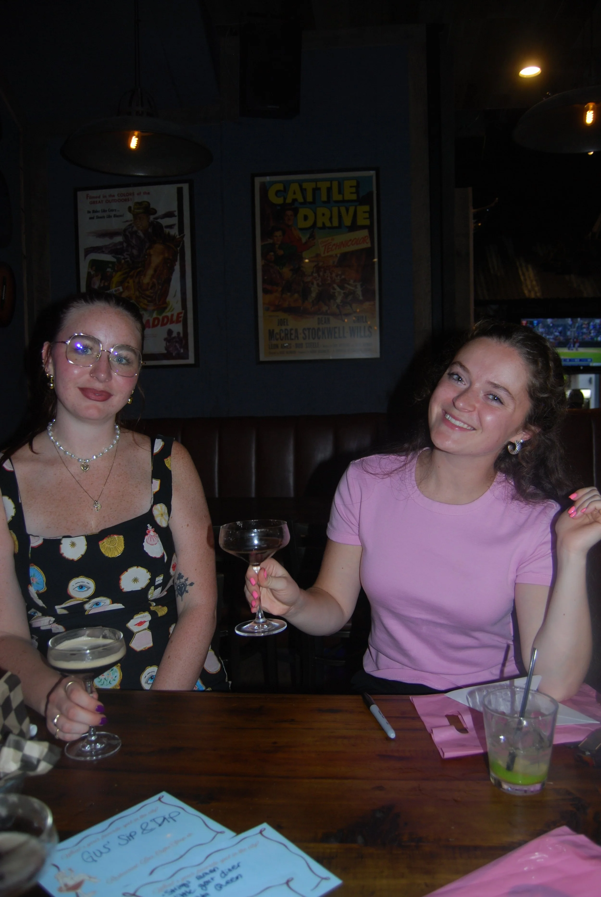 Two young women sitting at a table in a dimly lit restaurant, each holding a drink, smiling at the camera, with posters hanging on the wall behind them.