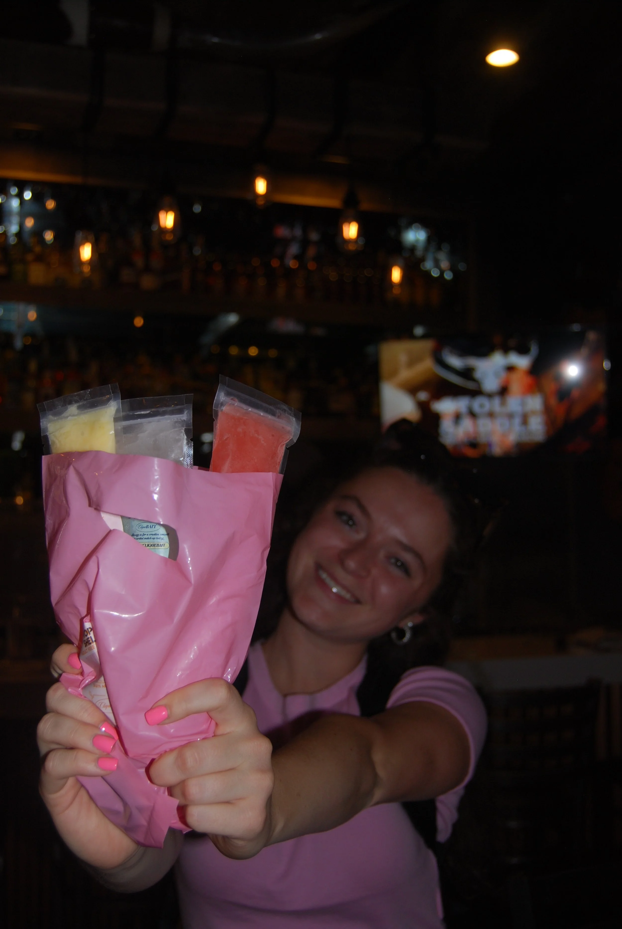 A smiling woman holding a pink paper bag with confetti shapes visible inside, in a dimly lit bar or restaurant setting.
