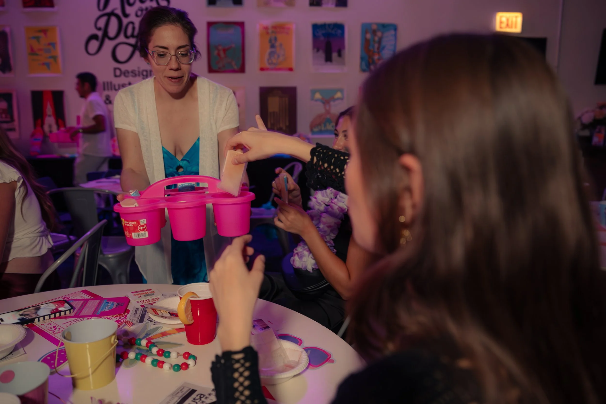 A young woman putting a piece of paper into a pink plastic bucket at a party with children and adults, surrounded by party decorations, colorful posters, and tableware.