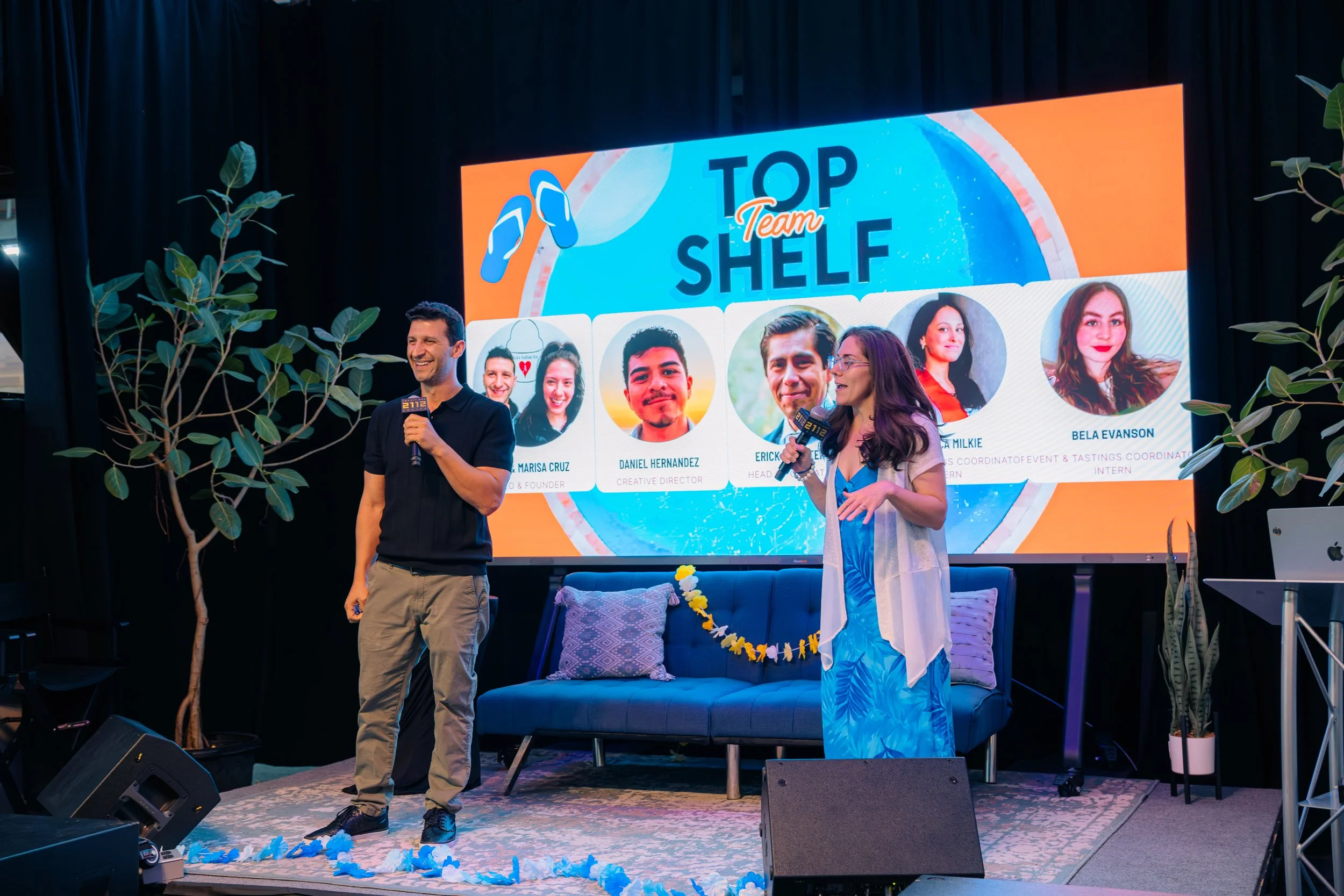 Two people speaking on stage in front of a large screen that reads 'TOP Team Shelf' with photos of team members. The man on the left holds a microphone and smiles, while the woman on the right also holds a microphone and gestures with her hand. There is a blue couch, potted plants, and a decorated stage area.