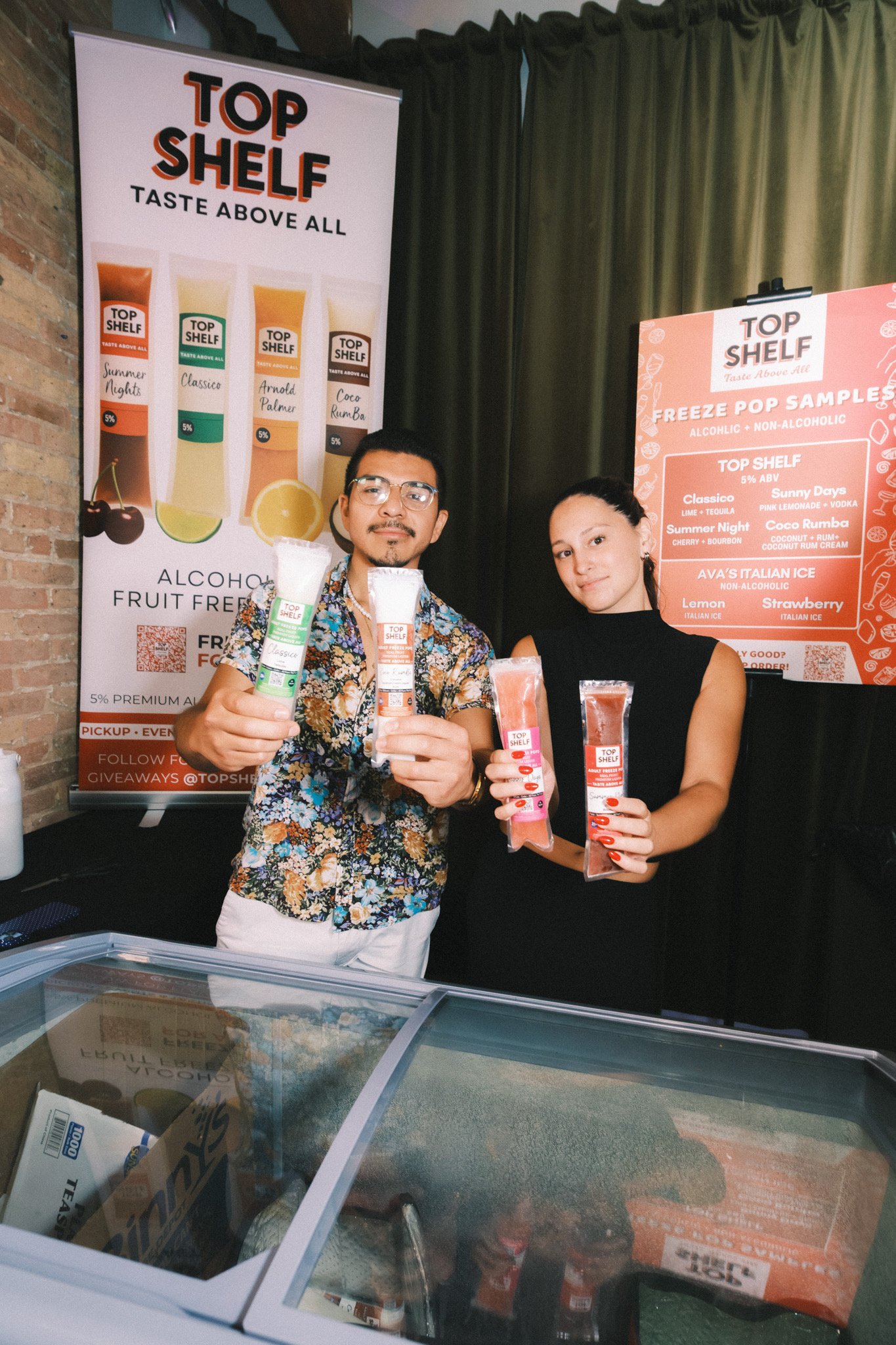 Two people holding frozen cocktail samples at a promotional event for Top Shelf, with banners displaying various drink flavors and options in the background.