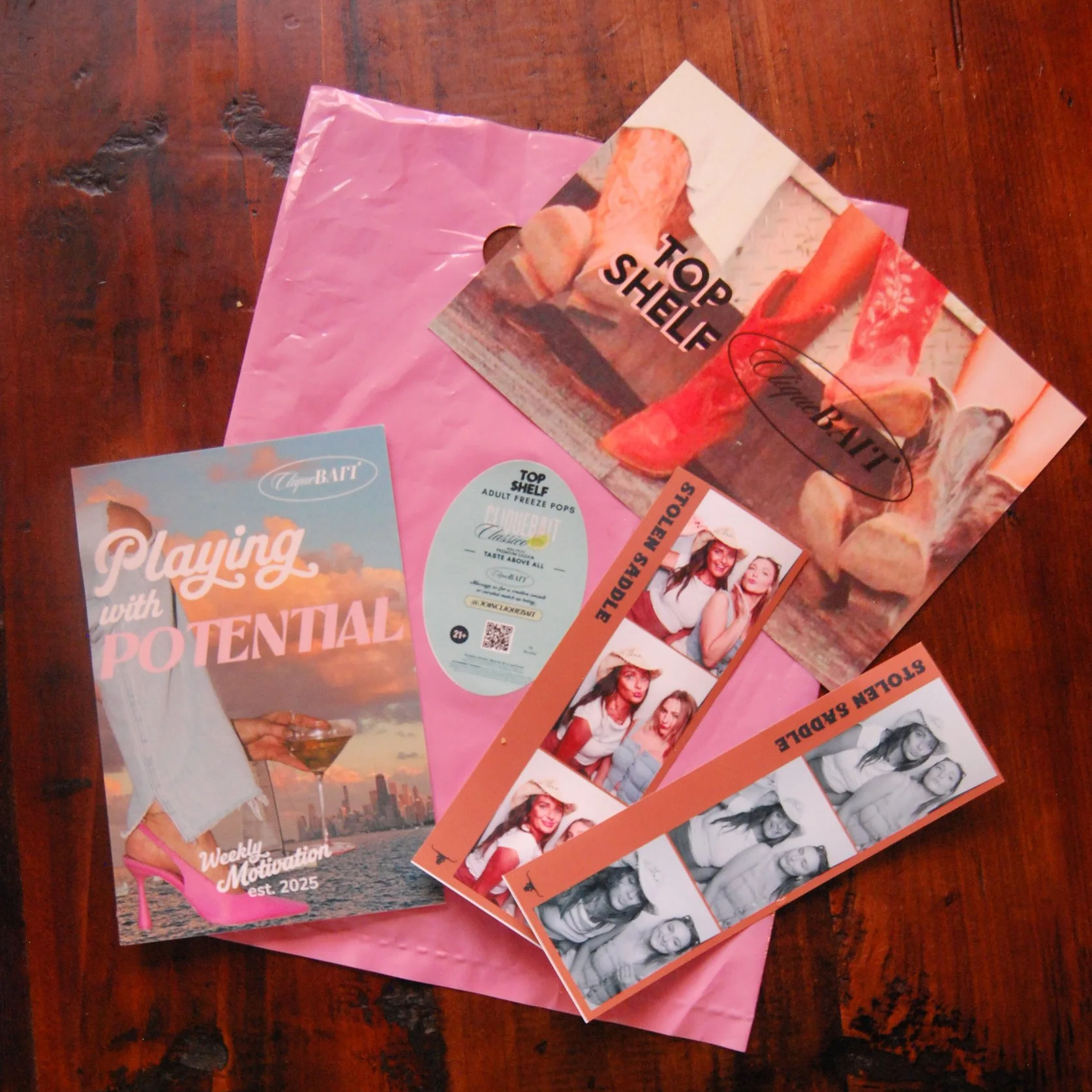 Promotional materials including flyers and photo booth strips with images of women, on a pink plastic bag on a wooden surface.