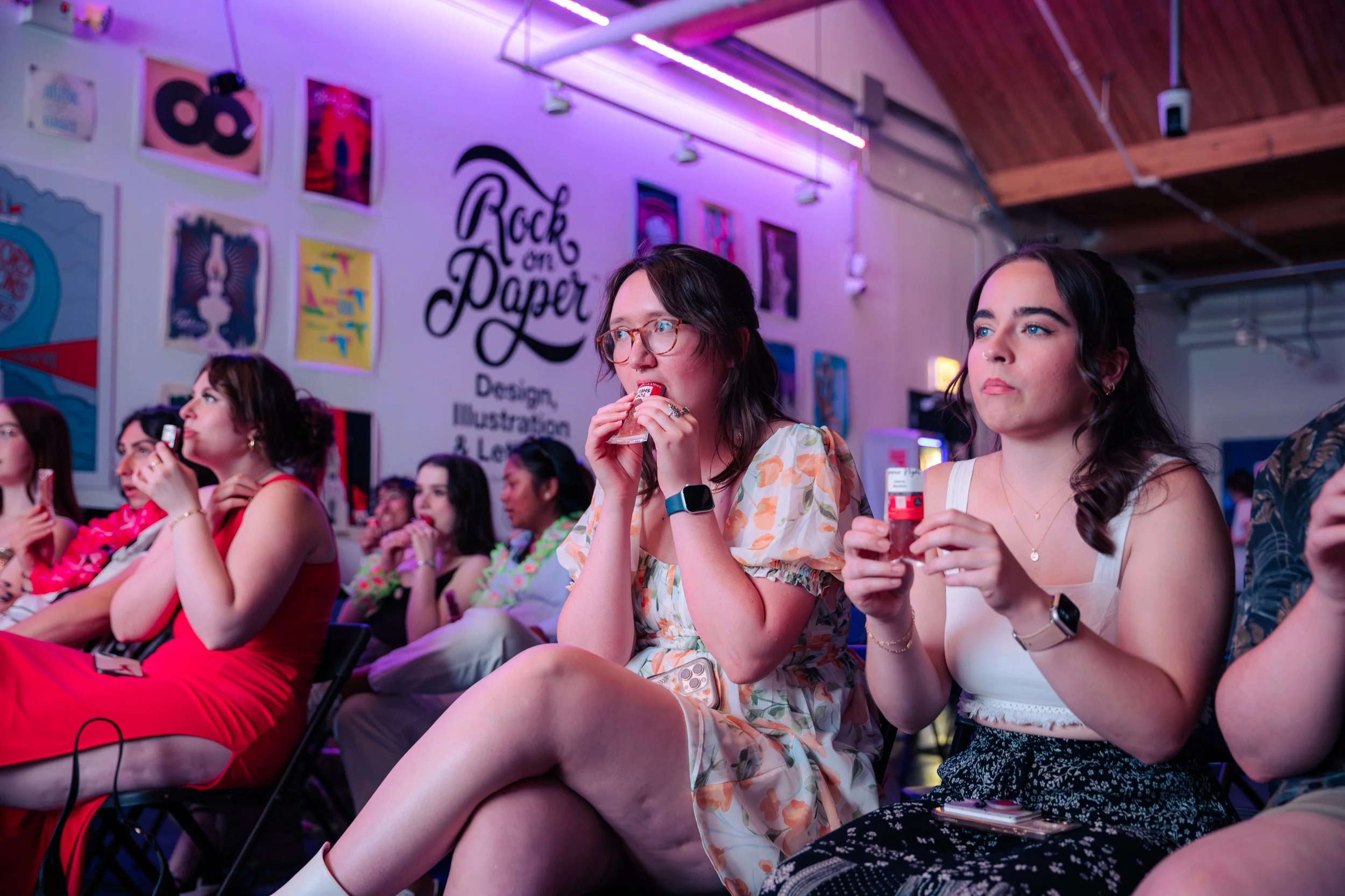 Group of women sitting in an audience at a design and illustration event, some eating snacks and wearing floral or colorful outfits, in a venue with art posters and a mural on the wall that says "Rock on Paper."