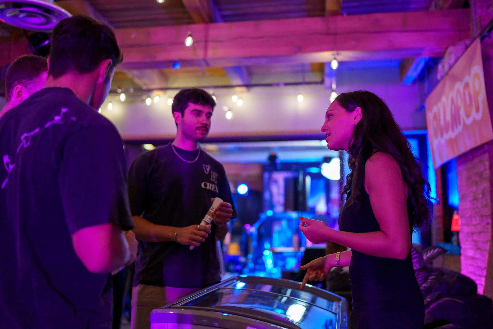 Four young adults are talking at a bar or nightclub. One woman is engaged in conversation with two men, while another man looks on. The setting has vibrant purple and blue lighting, with drums and musical instruments visible in the background.