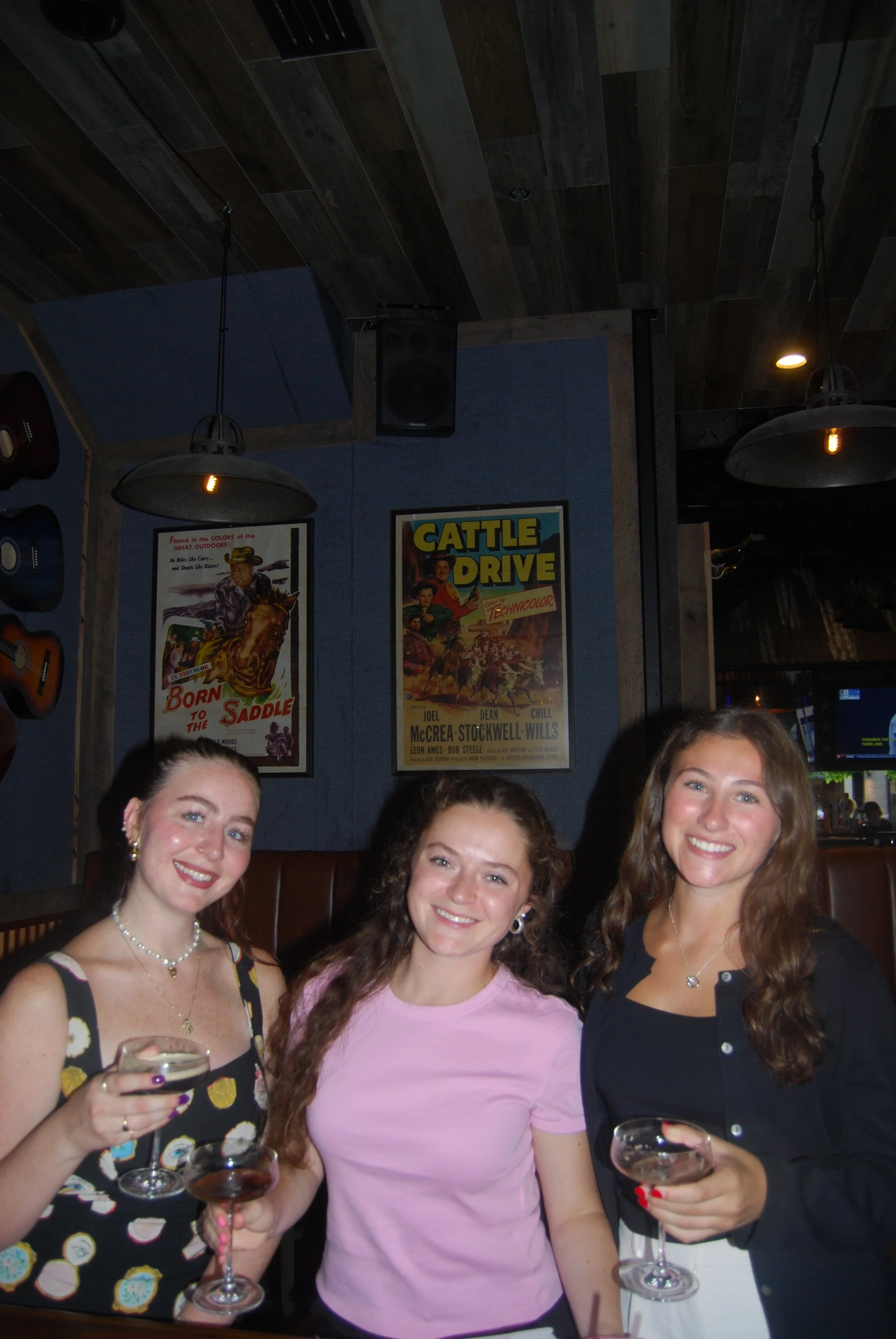 Three young women smiling and holding drinks at a restaurant or bar, with vintage posters and guitars on the wall behind them.