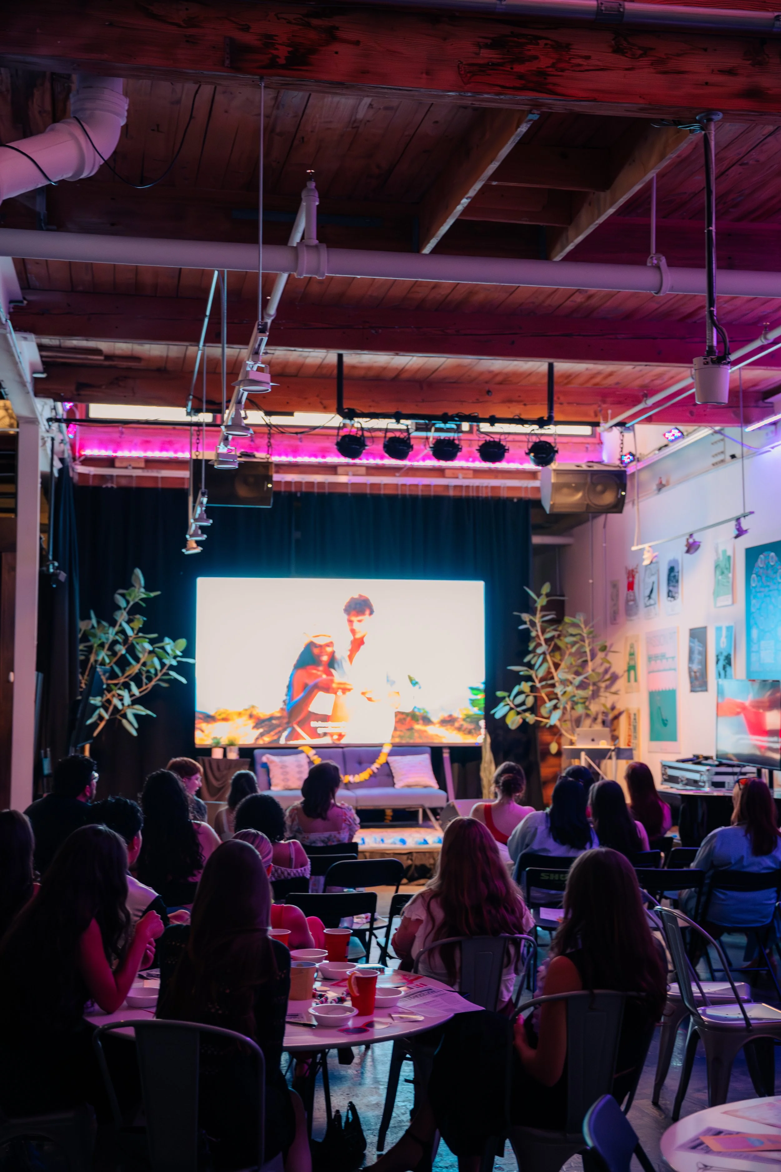 Indoor event space with audience watching a large screen showing a woman and a man, with plants and art on the walls, and tables set with drinks.