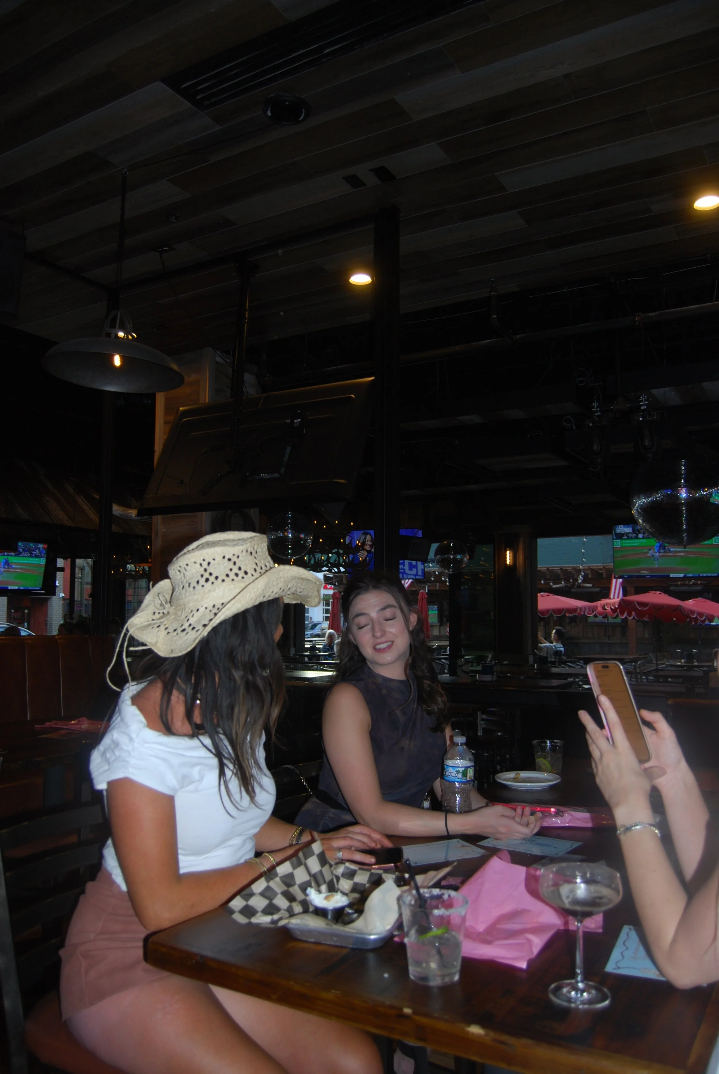 Two women sitting at a table in a restaurant or bar, one wearing a white top and pink shorts with a straw cowboy hat, the other in a dark sleeveless top, with drinks and food on the table, and a person taking a photo with a smartphone.