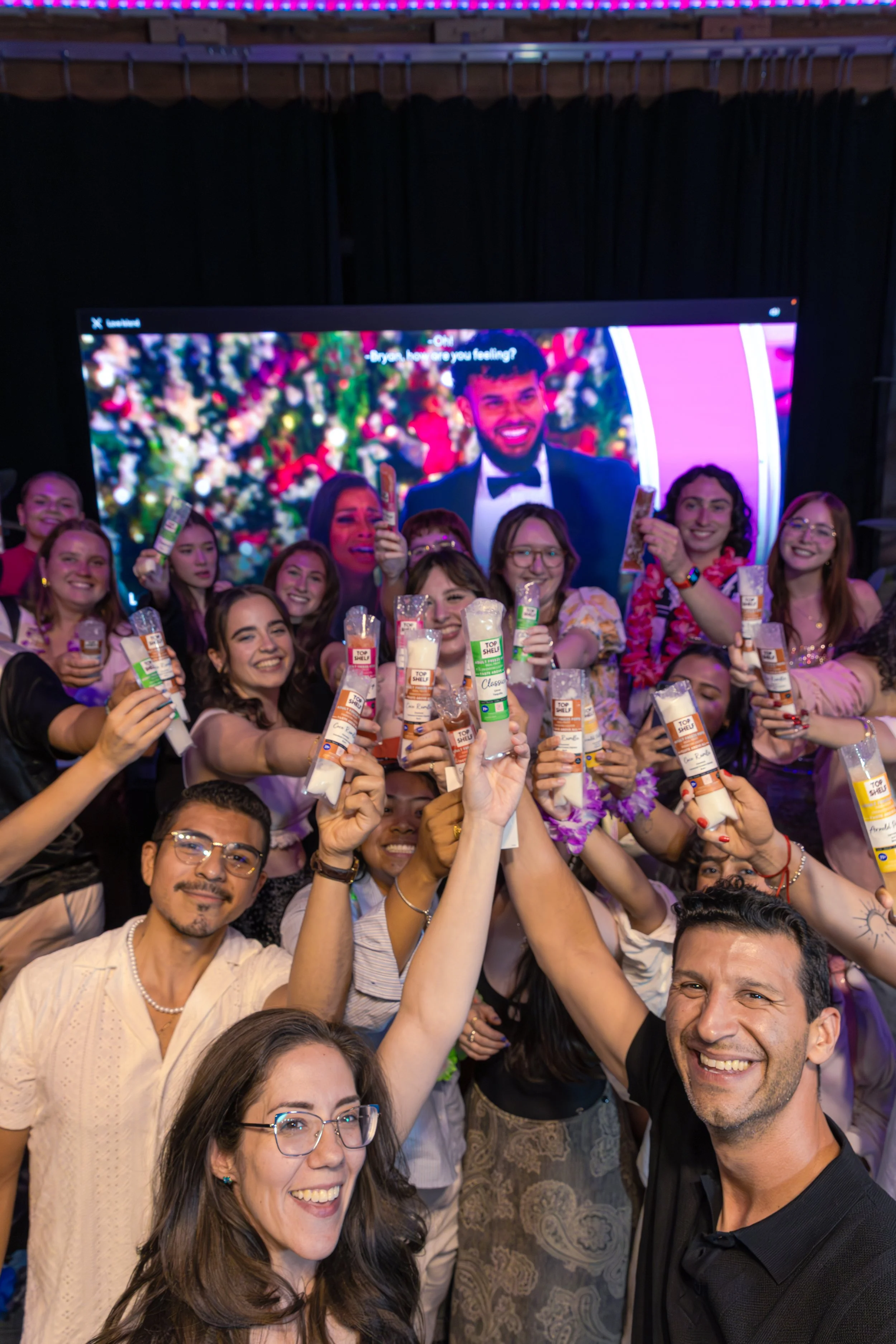 A group of people at a celebration, holding snacks and smiling, with a large screen in the background displaying a man in a tuxedo and woman at a Christmas-themed event.