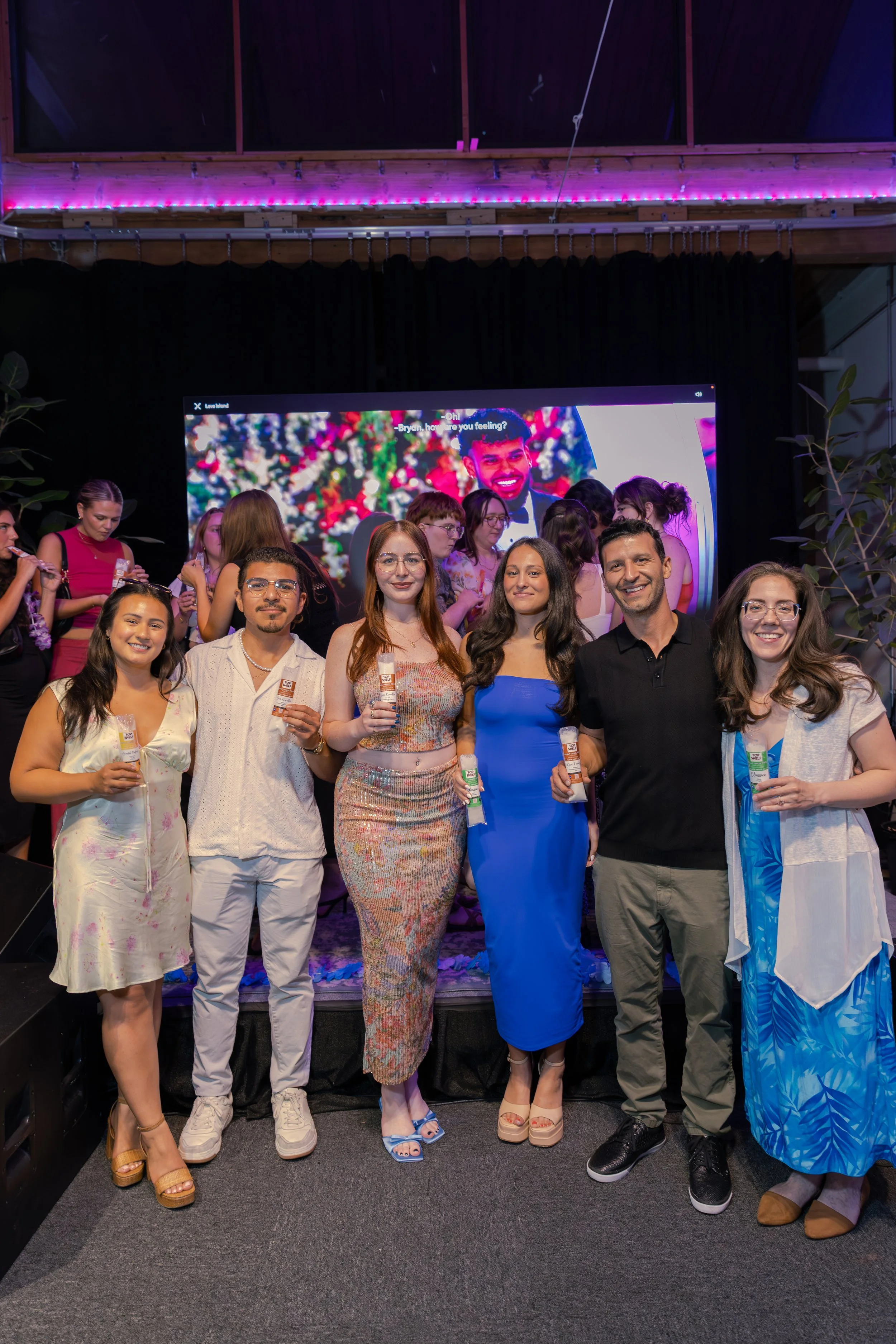 Group of six people standing on stage with drinks, smiling at camera at a party or celebration, with a large screen and other people in the background.