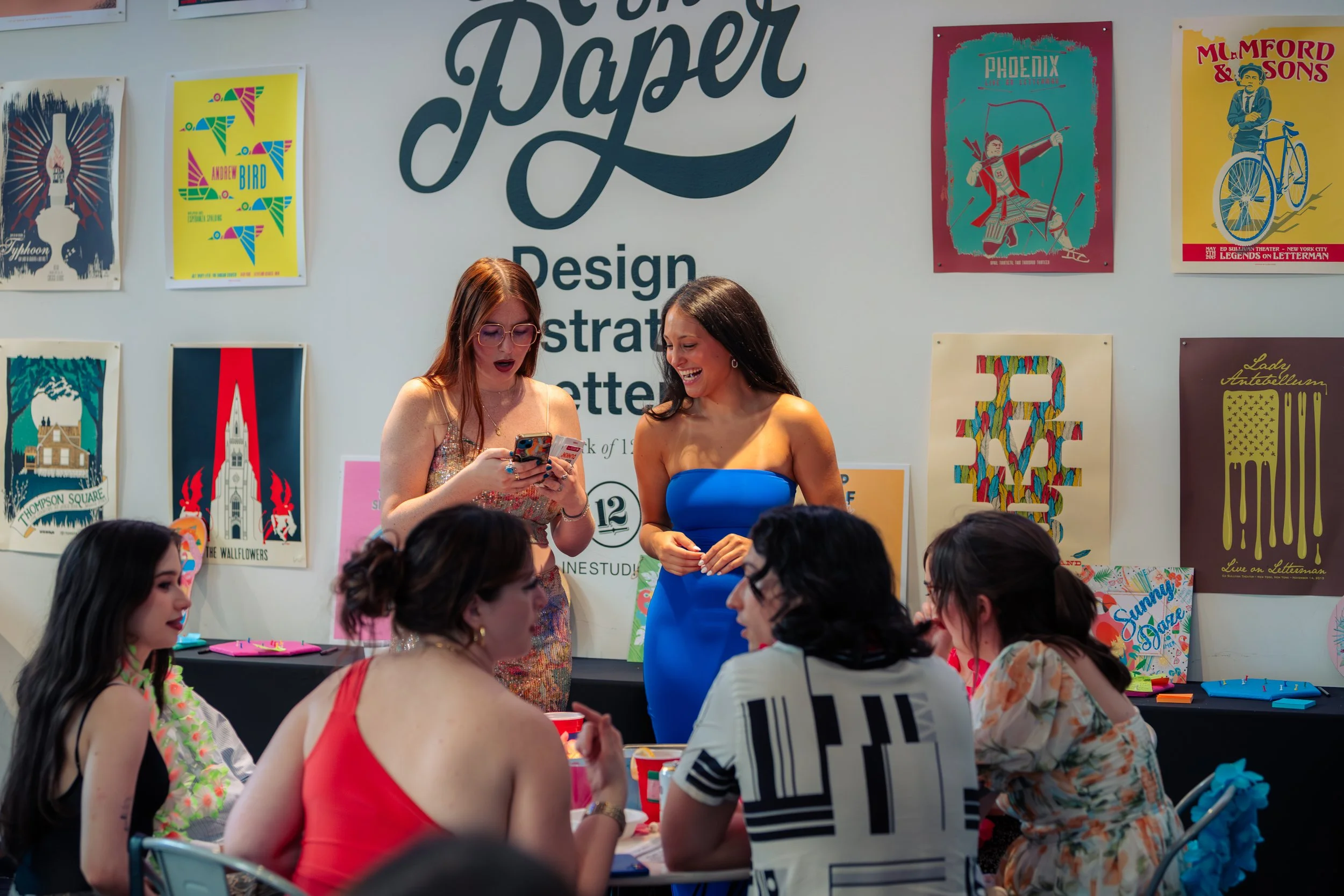Group of women at a social gathering in an art studio or gallery, with colorful posters on the wall and people around a table.
