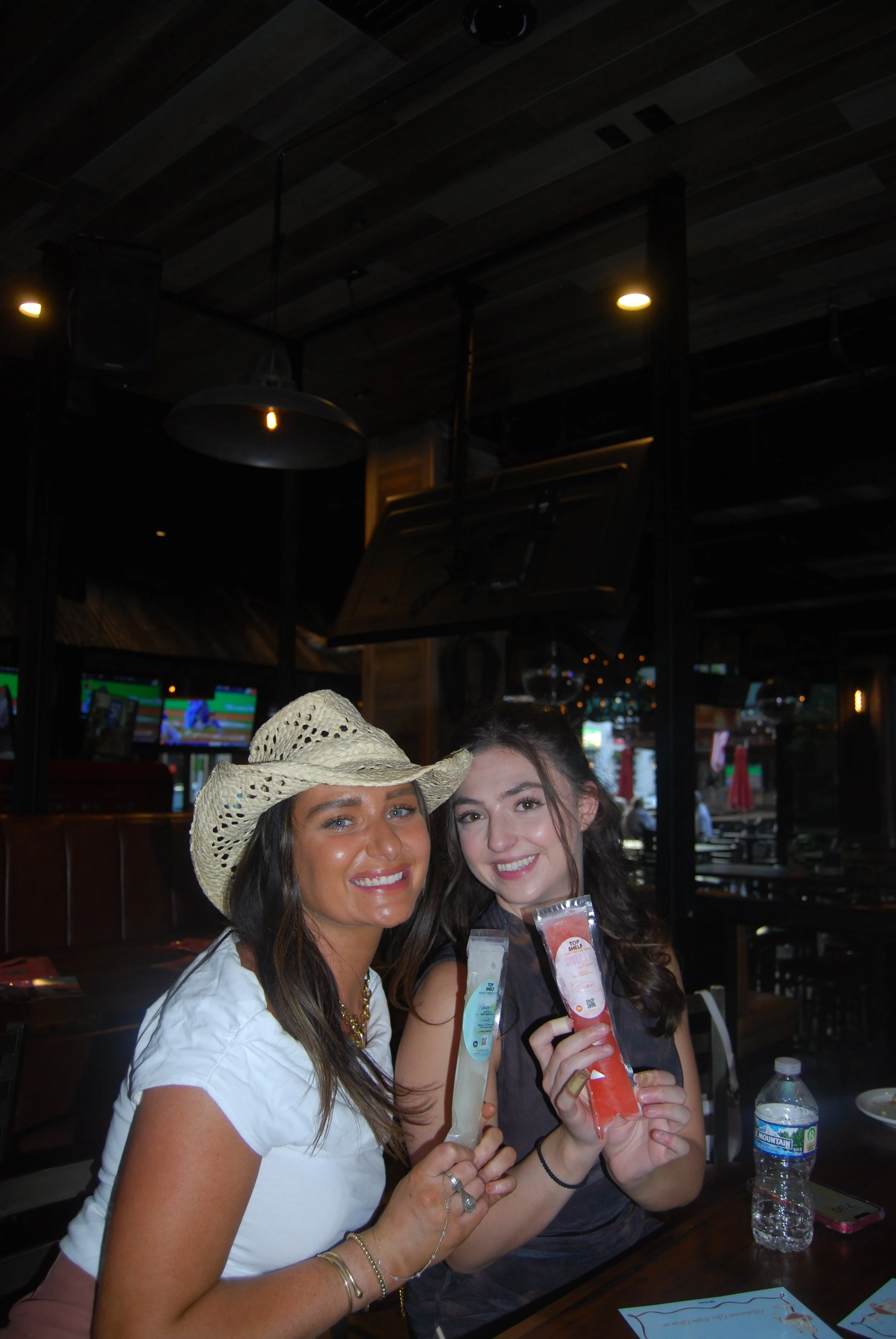 Two women smiling and holding colorful drinks in a dimly lit restaurant or bar.