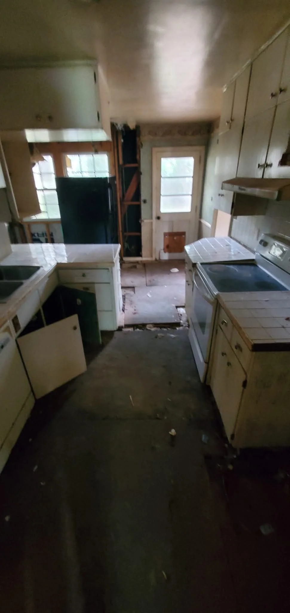 A dilapidated kitchen with missing cabinet doors, a stained floor, and a cluttered countertop, leading to a door outside with a window and a pet door at the bottom.