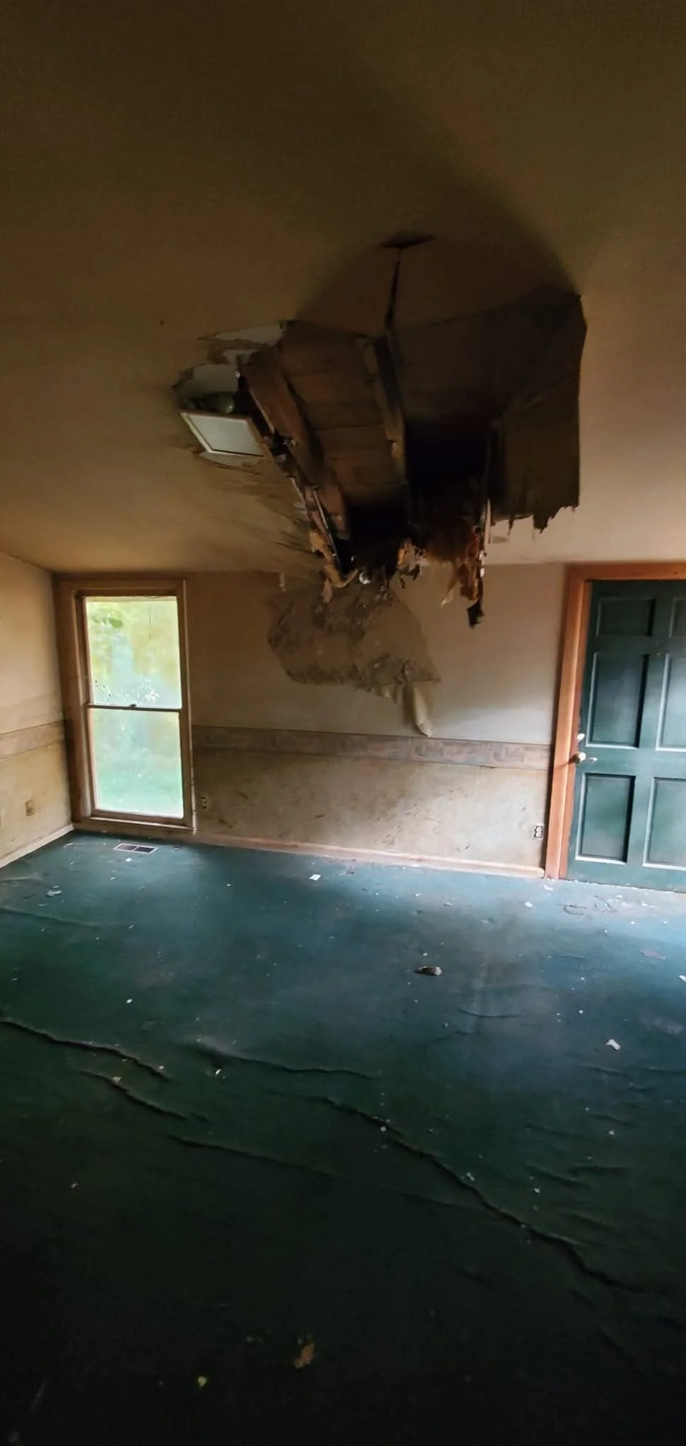 A room with a damaged ceiling hanging down, possibly from water damage or structural failure, with a green carpeted floor, a window with natural light coming in, and a closed dark green door.