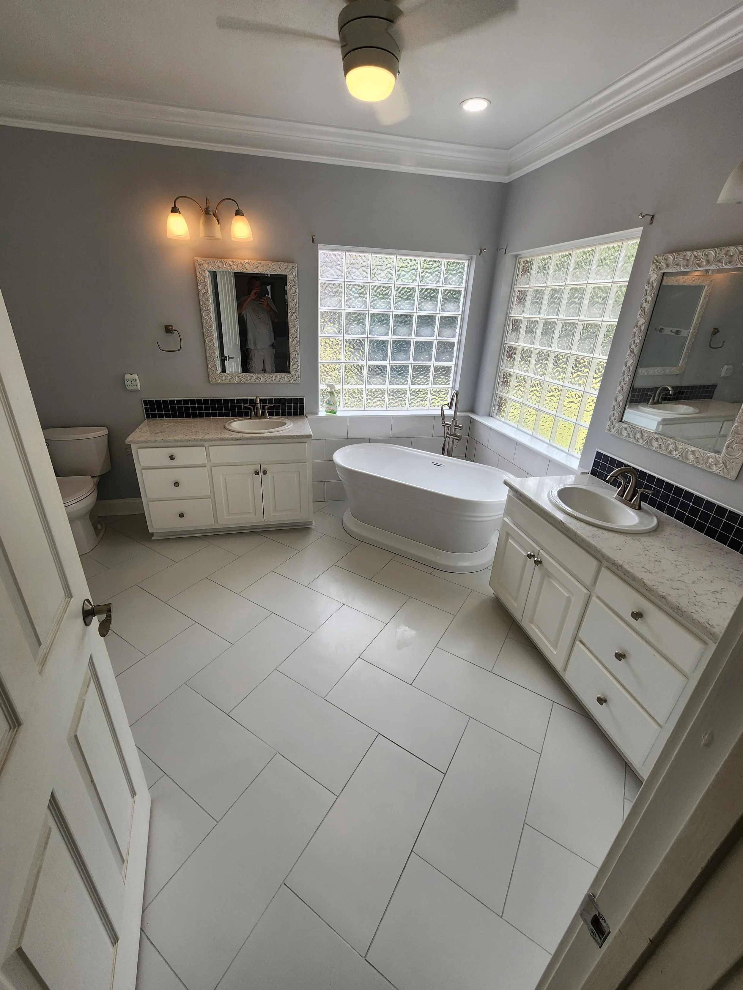 Bathroom with a bathtub, two sinks with white cabinets, a toilet, two mirrors, two glass block windows, a ceiling fan, and lighting fixtures.