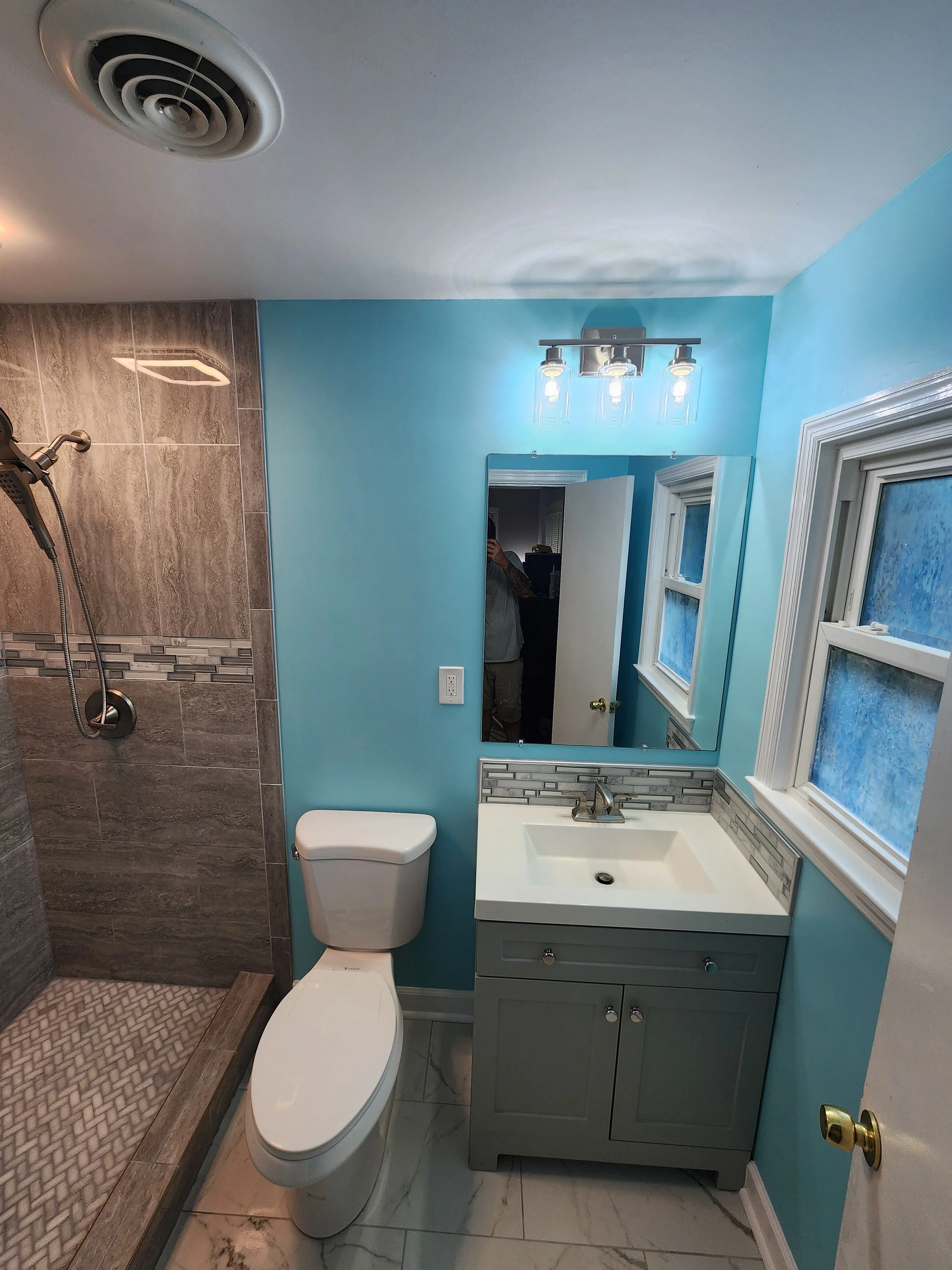 Bathroom with a shower area on the left, a toilet in the center, and a vanity with a sink and mirror on the right. There are two windows above the vanity and a light fixture with three bulbs above the mirror.