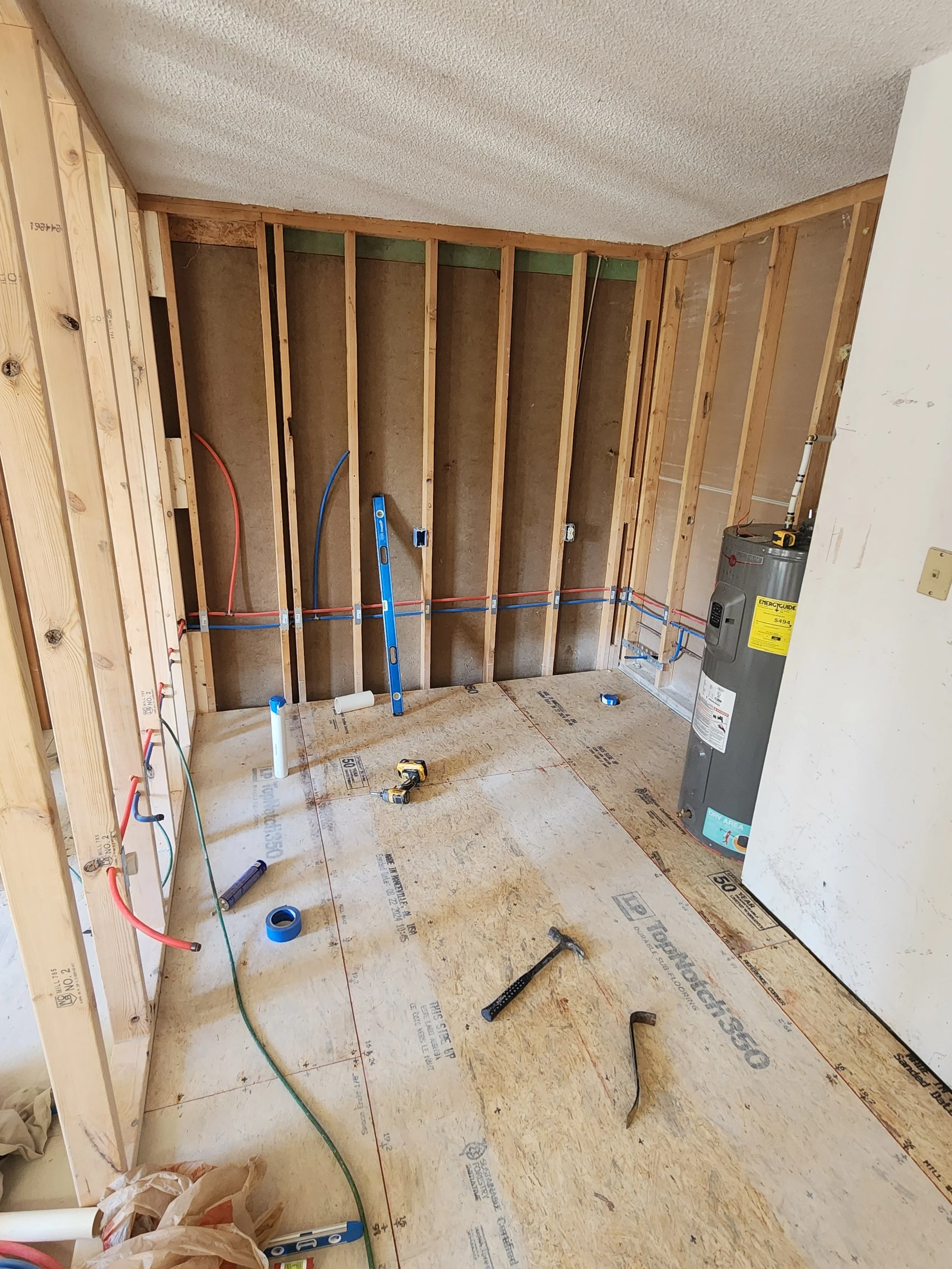 Room under construction with exposed wooden wall studs, electrical wiring, and plumbing pipes, and a water heater.