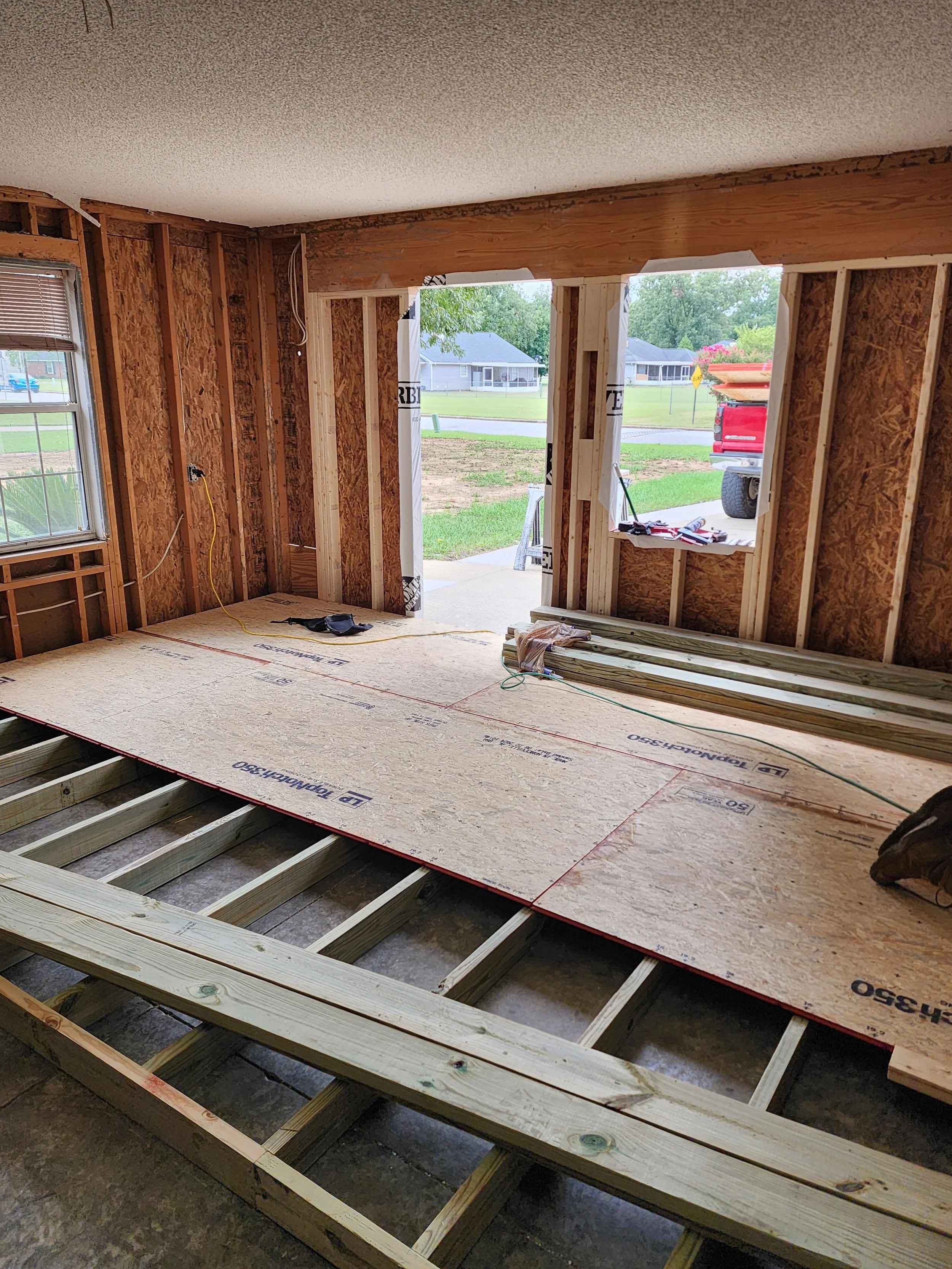 Interior of a house under construction, showing exposed wooden framing, partially installed subfloor, and an open doorway leading outside, with construction tools and materials scattered around.