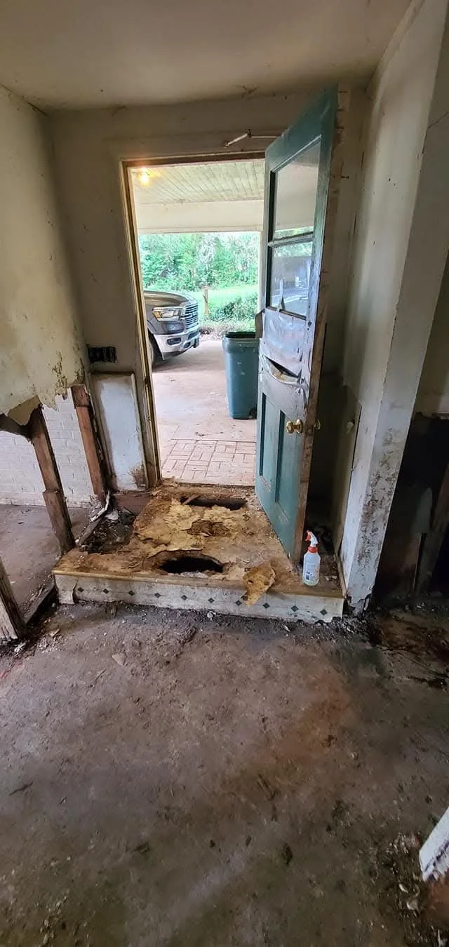 Interior view of a house with a damaged doorway, showing exposed wall studs, a partially removed floor with two large holes, and cleaning supplies on the ground.