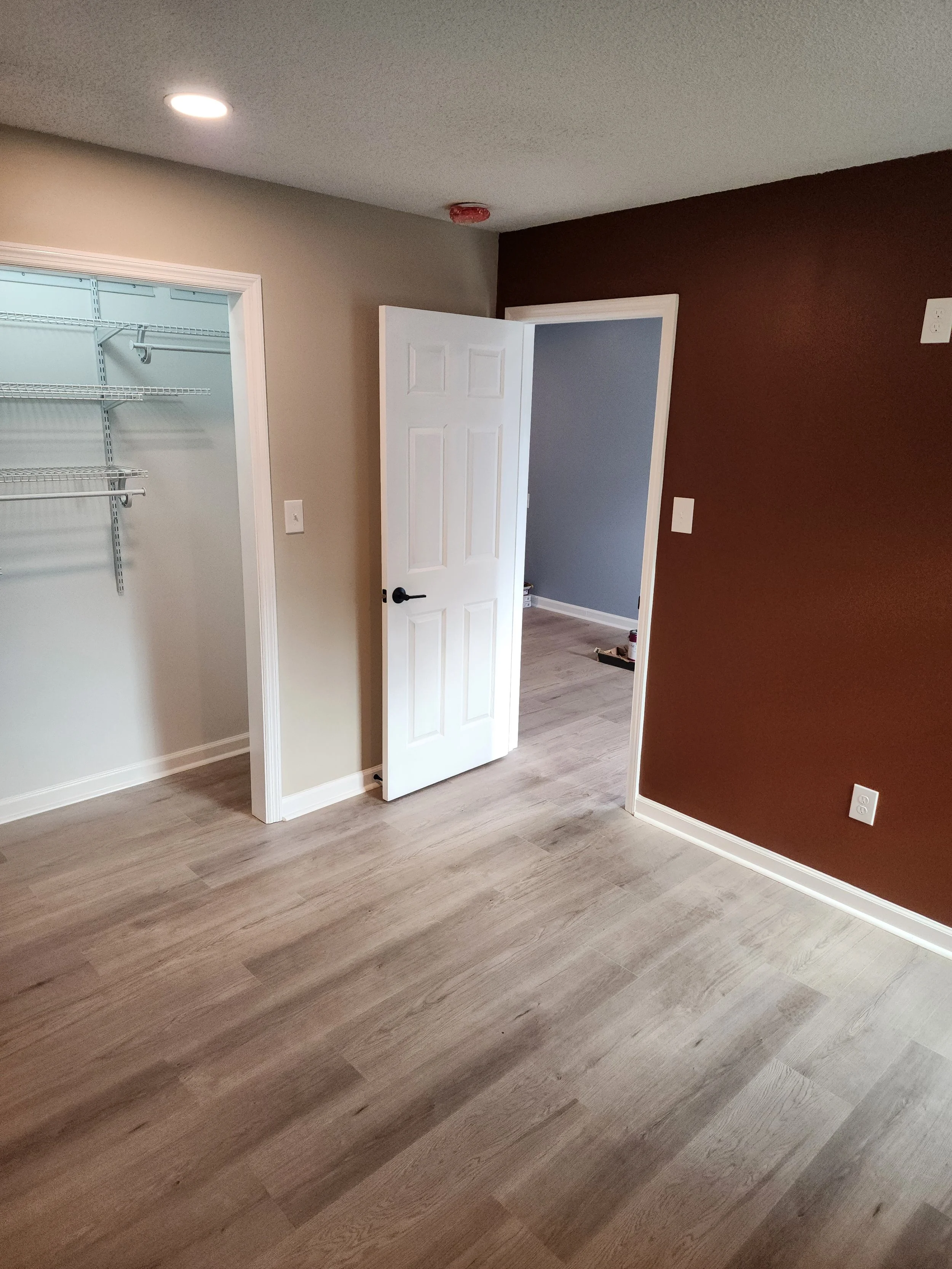 Empty room showing open closet with wire shelves, beige and rust-colored walls, white door, light wood flooring, and visible electrical outlets.