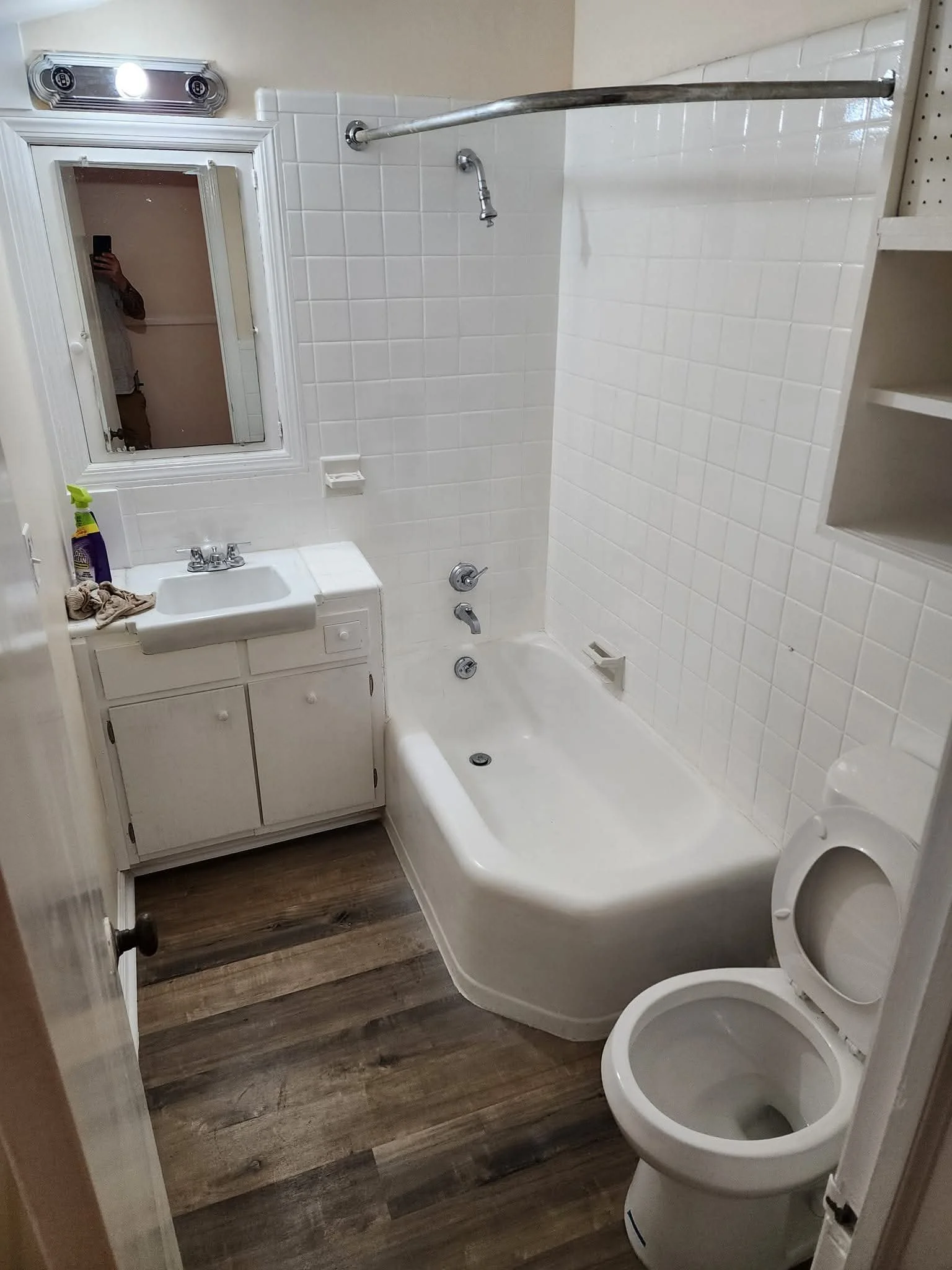 Small bathroom with a bathtub, toilet, small sink with cabinet, mirror, and storage shelves. The bathroom has wood-look flooring and white tiled walls.