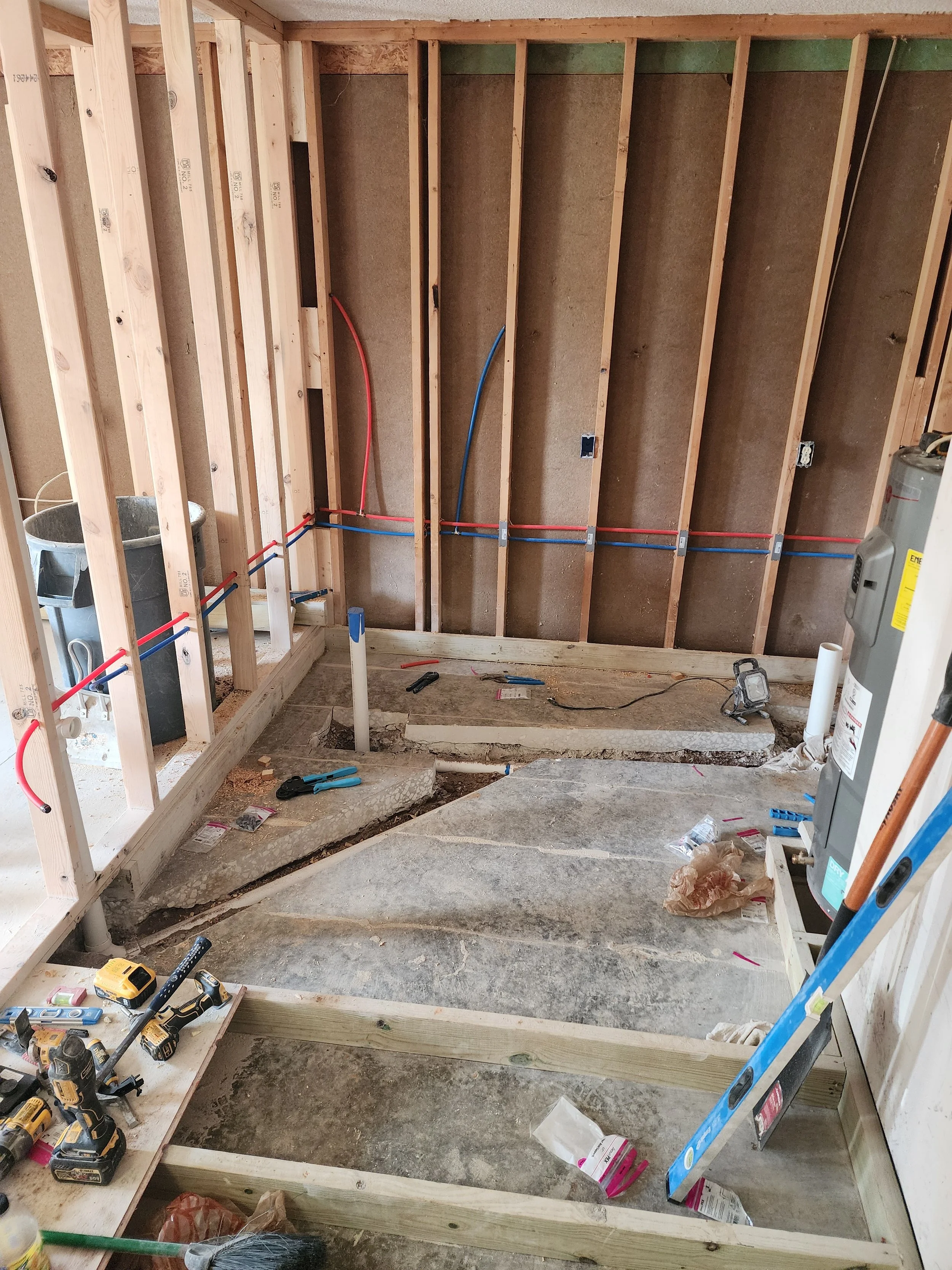 Construction site showing partially built wooden frame walls, exposed electrical wiring, tools, and construction materials on the floor, with the subfloor partially removed.