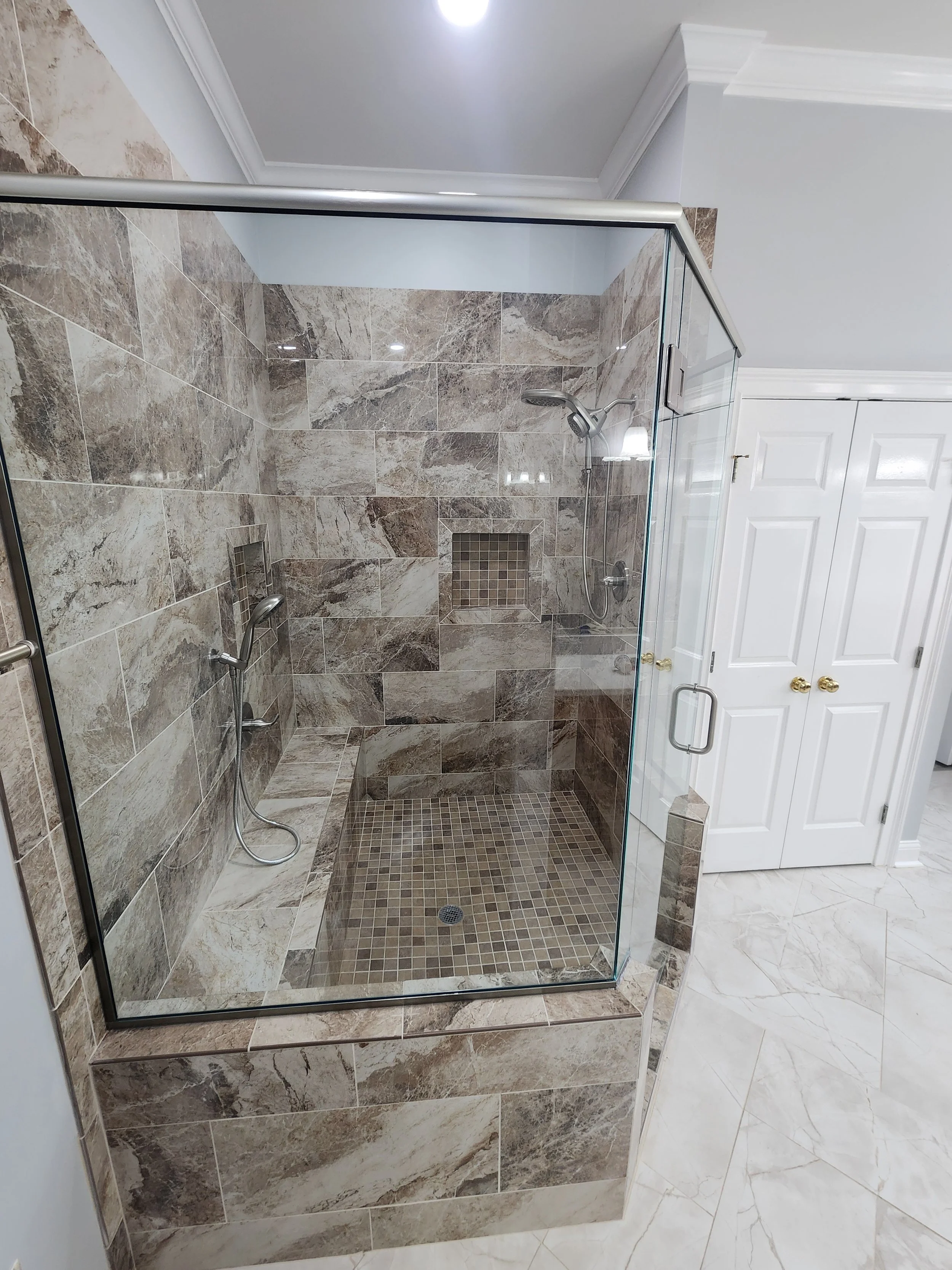 A glass-enclosed walk-in shower with marble tile walls and mosaic tile floor, featuring two adjustable showerheads and a built-in niche.
