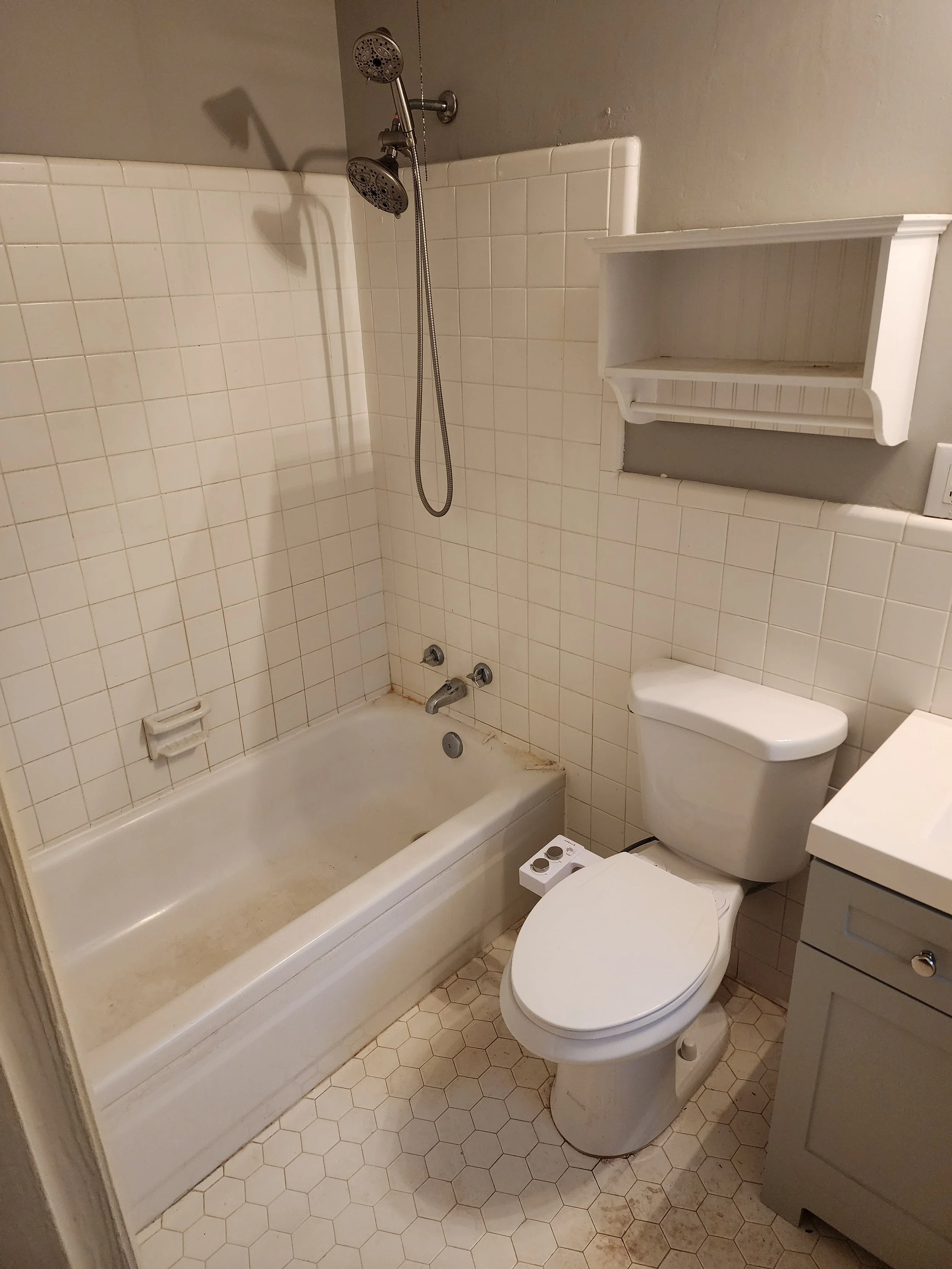 Small bathroom with bathtub, toilet, and wall-mounted shelf. The bathtub appears dirty with stains. The floor tiles are hexagonal, and the wall tiles are square. The mirror and sink are not visible in the image.