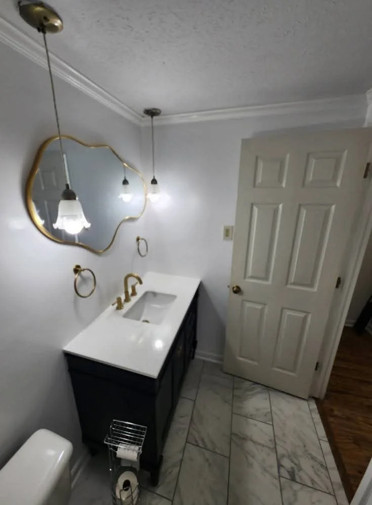Bathroom with a white countertop vanity, gold accents, a mirror with a gold frame, and wall-mounted gold towel rings. The bathroom has white walls, marble tile flooring, and a white door.