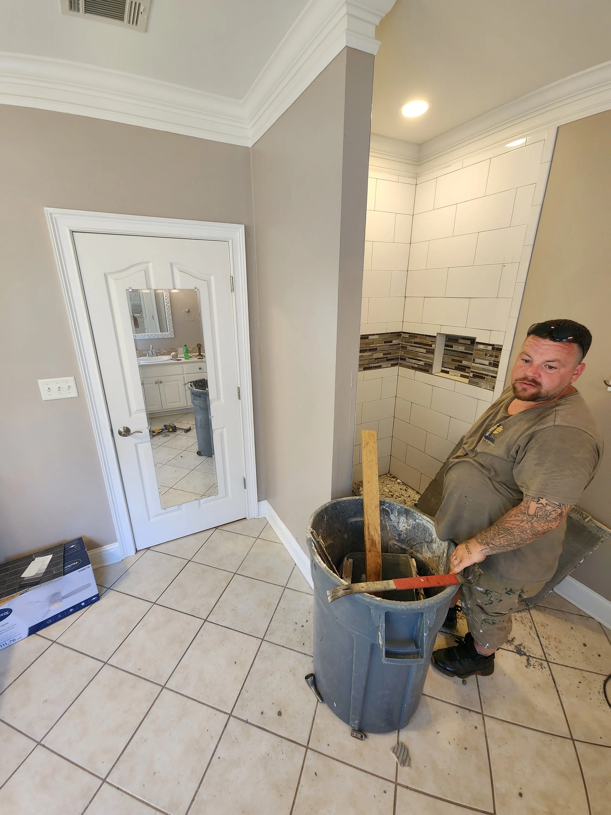 Man working on a bathroom renovation, standing next to a large trash bin with tools, in a room with beige tiles and a—partially finished—shower with decorative tile accents.