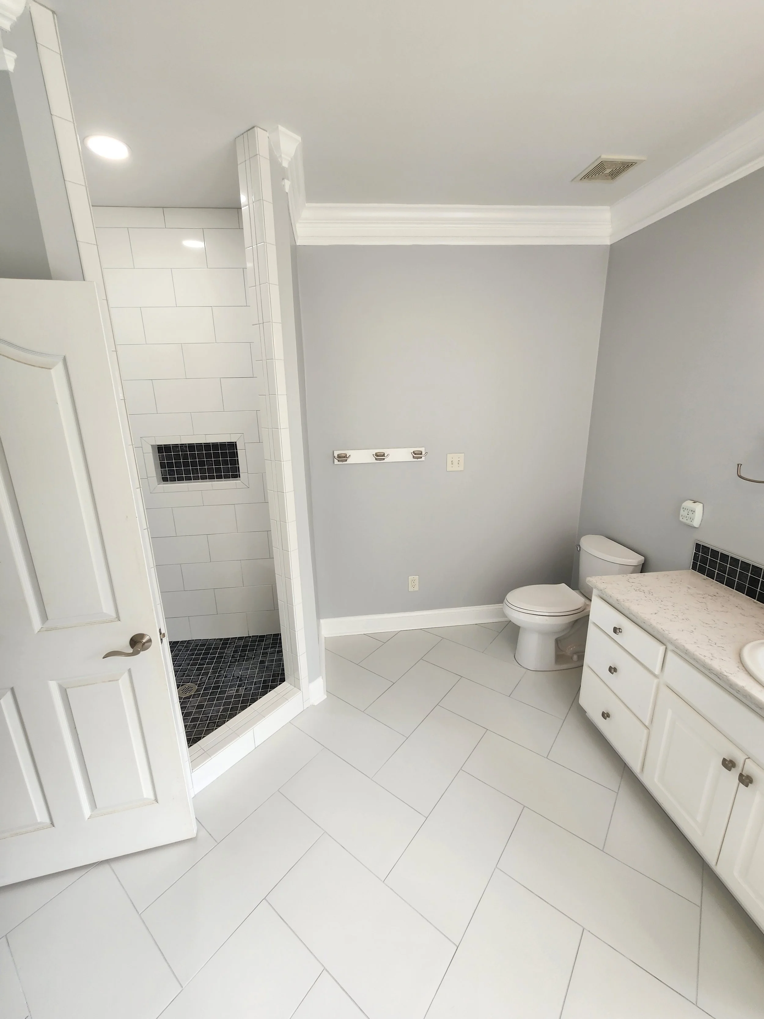 A modern bathroom with white tiled floors, a shower stall with white tiles and black accents, a white cabinet with a marble top, a toilet, and light gray walls.