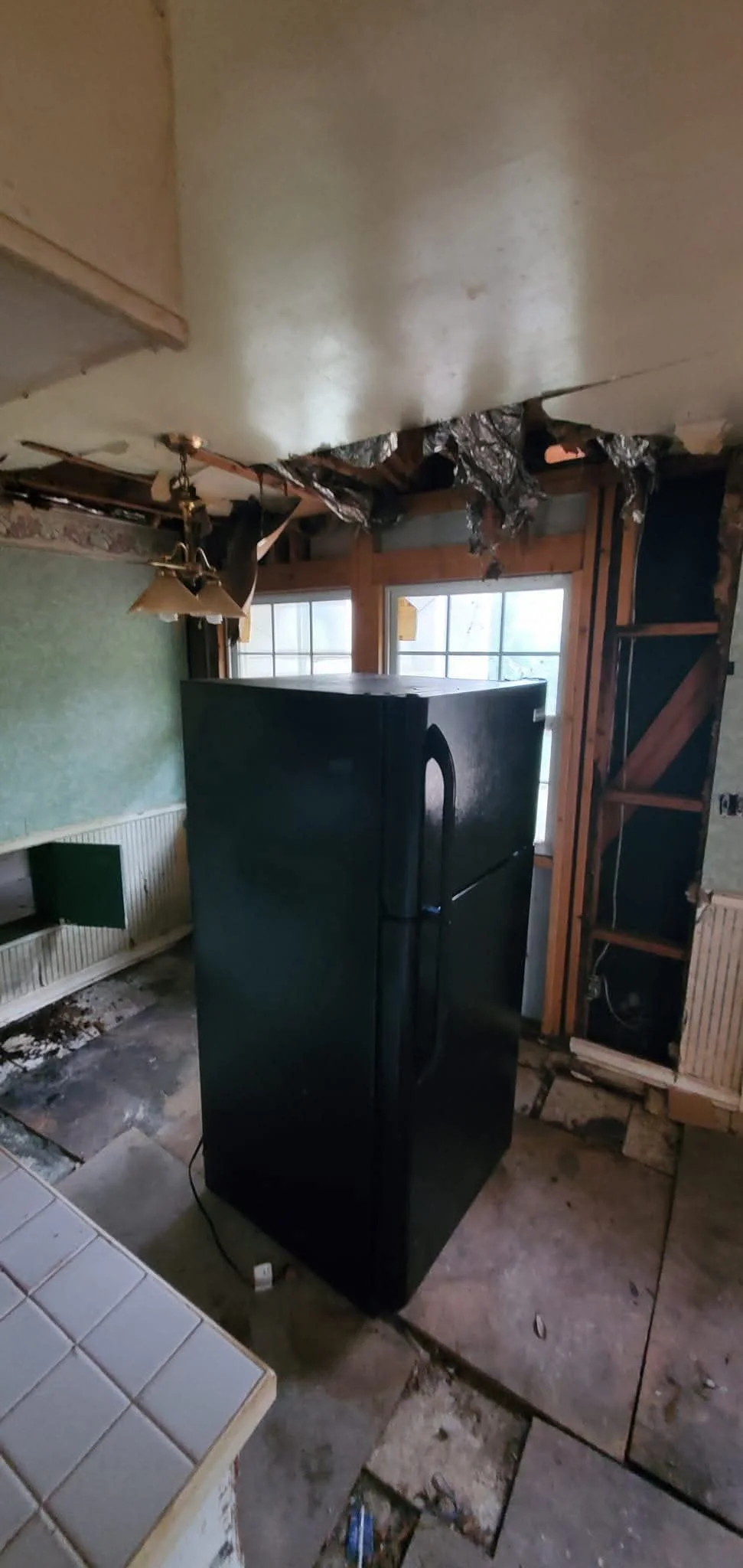 The image shows a black refrigerator in a room under renovation, with exposed wooden framing, damaged ceiling, and partially removed wall paneling. There is a window in the background and construction debris on the floor.