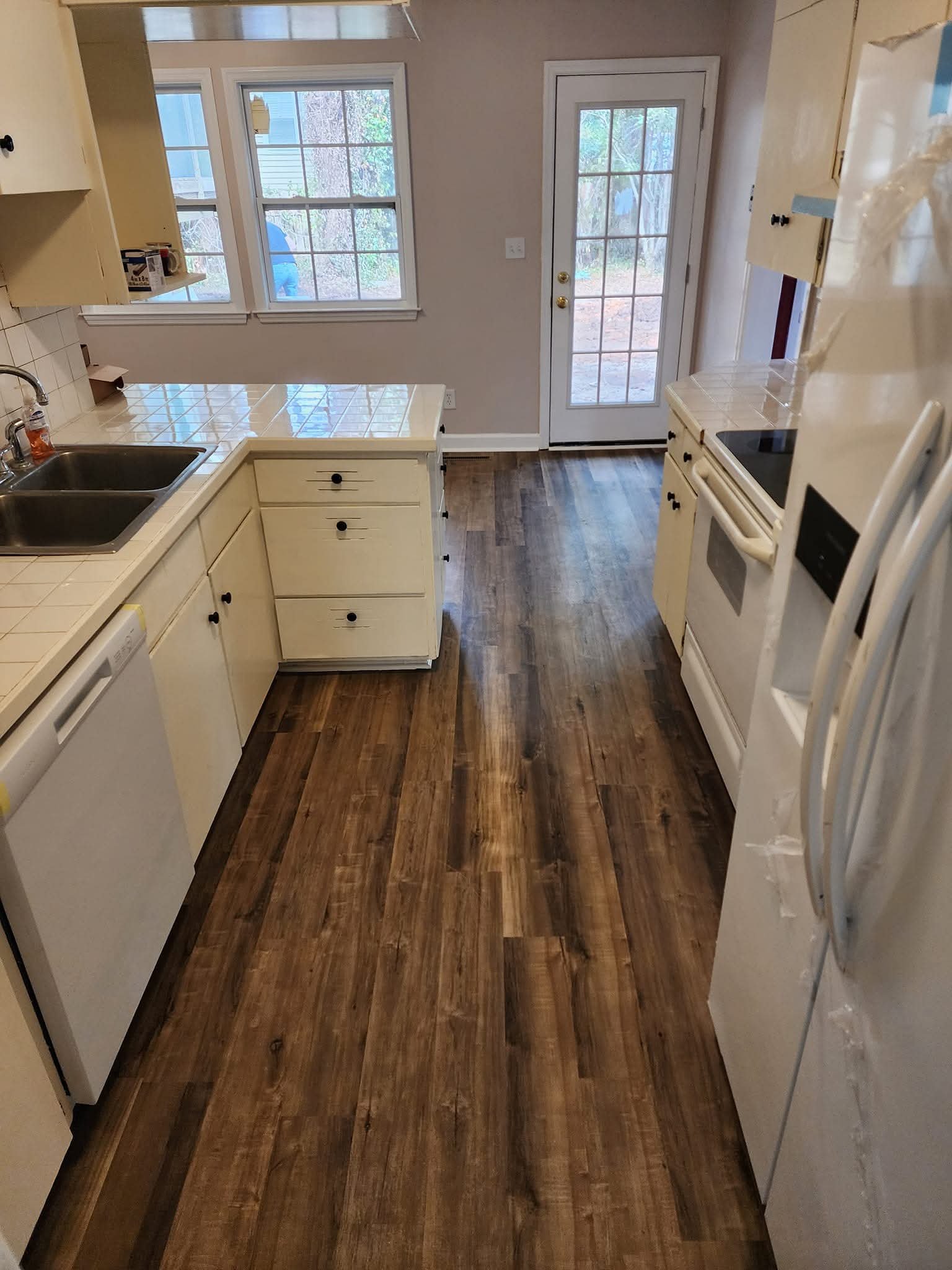 Kitchen with wooden floors, white cabinets, double sink, refrigerator, stove, and a door with glass panes leading outside. Two windows are visible above the sink.