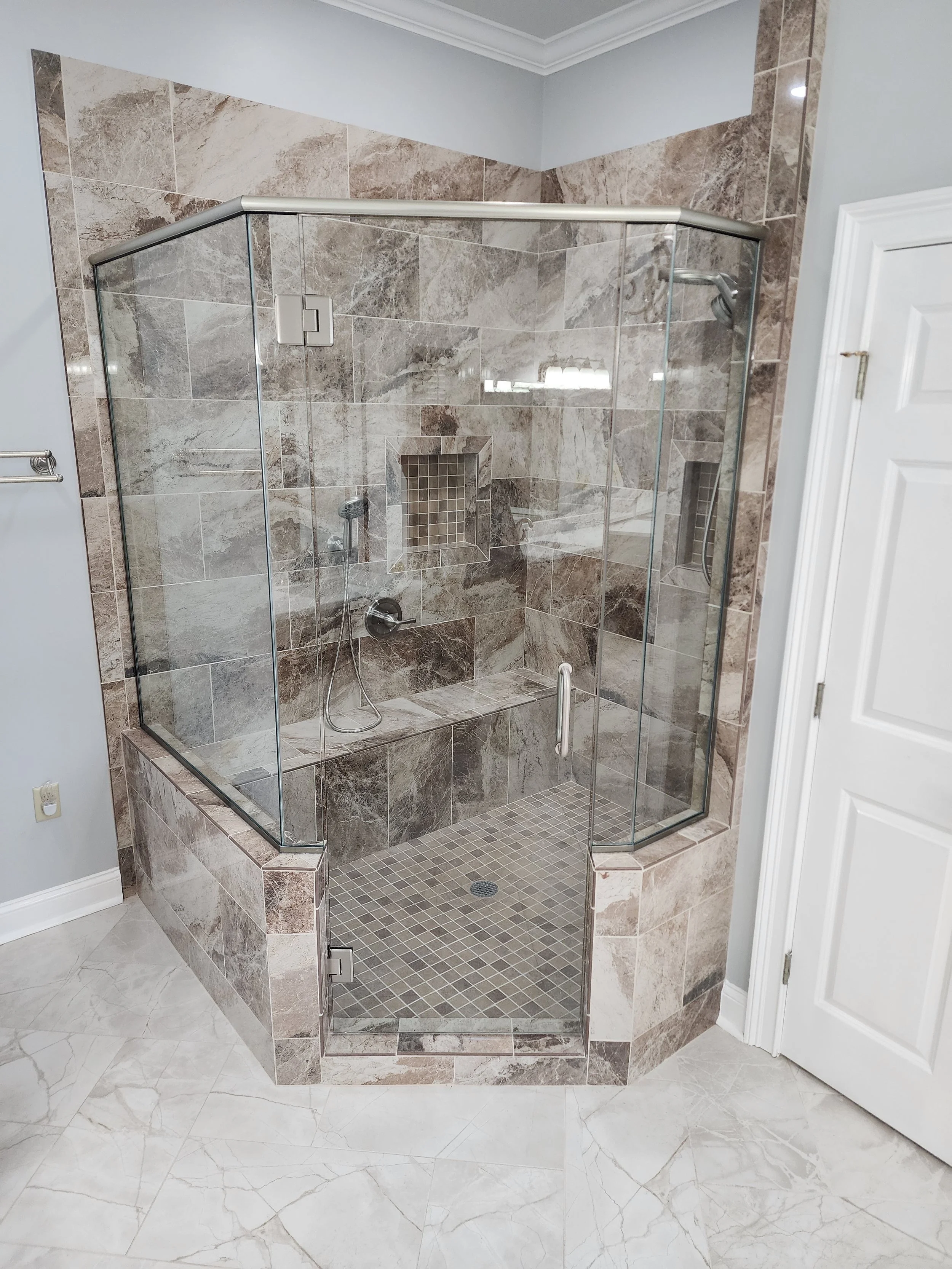A glass-enclosed walk-in shower with beige and brown marble tiles, built-in niche shelves, a bench, and a handheld showerhead, with a white door beside it.