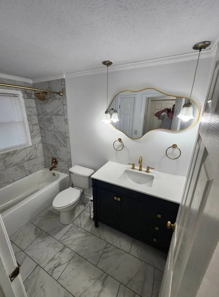 A small bathroom with a white bathtub and marble tiles on the wall and floor, a toilet, and a black vanity with a white countertop. Above the vanity, there is a wavy mirror, and two pendant lights hanging from the ceiling. The bathroom also has gold 