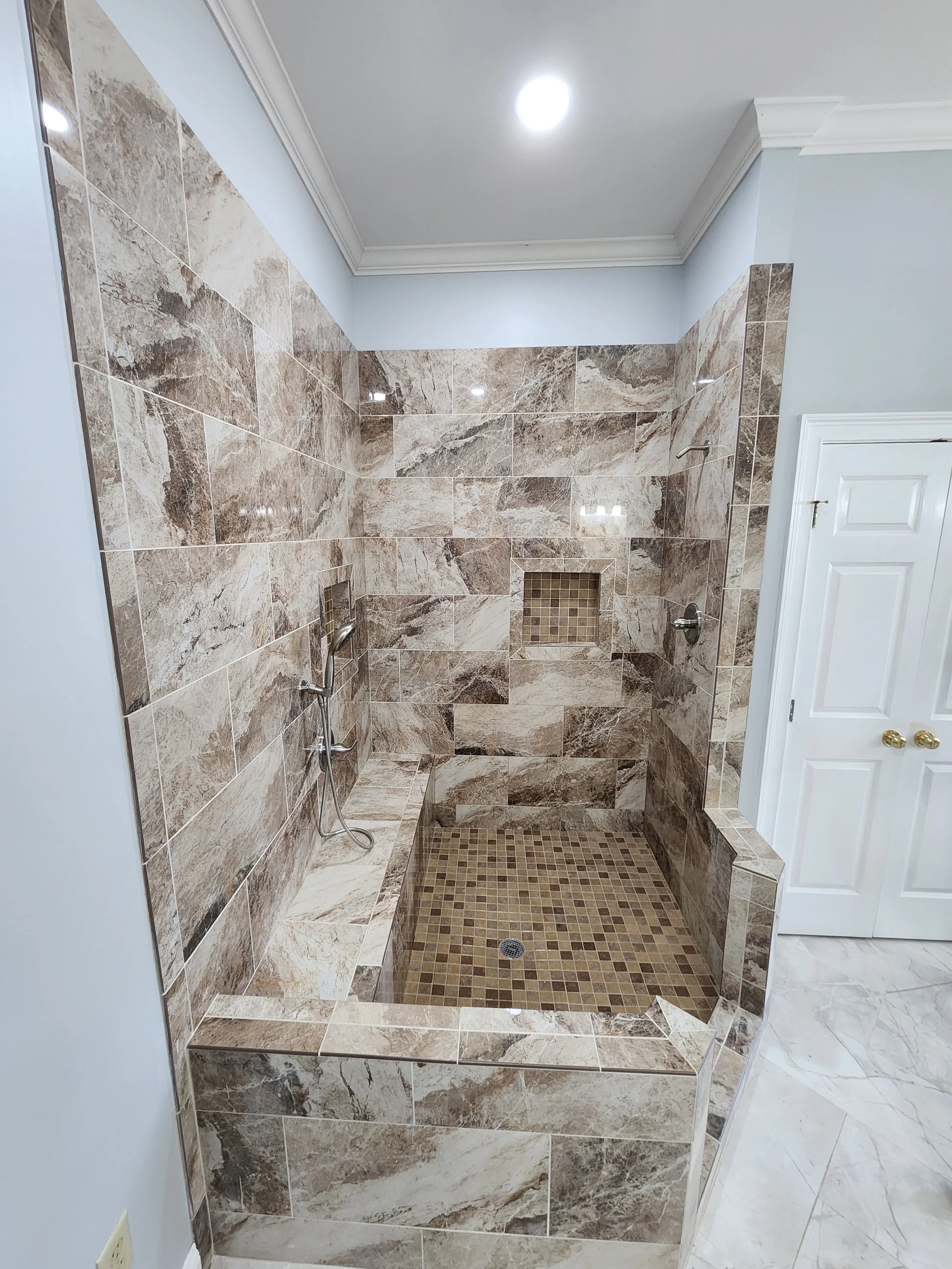 A walk-in shower with beige and brown marble tiles, a built-in bench, a small tiled niche, and a rain shower head. The shower has a partial enclosure and is located in a bathroom with light blue walls and white crown molding.
