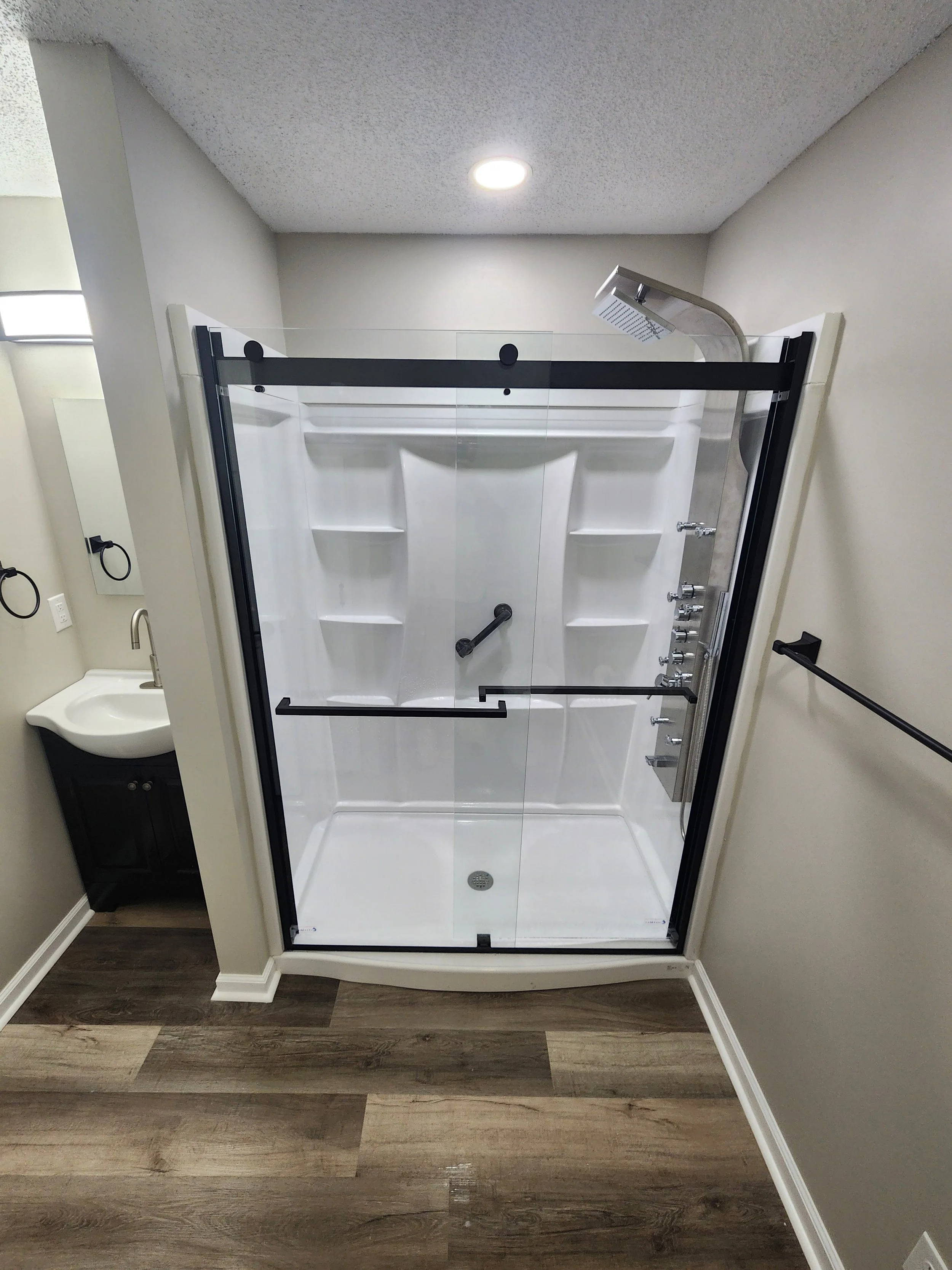 A walk-in shower with glass doors, a rain shower head, multiple body jets, and a grab bar, next to a small sink with a black cabinet.