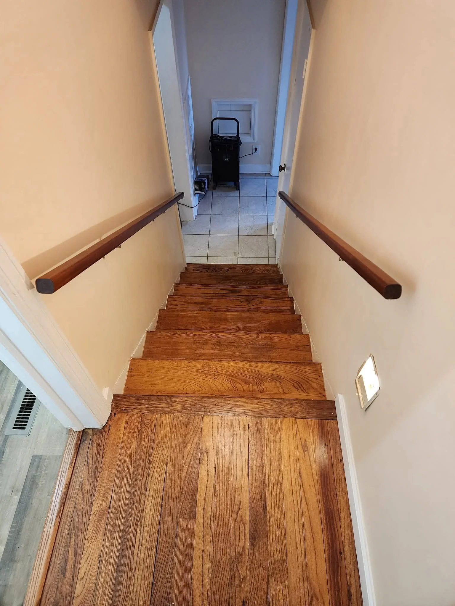 View of a staircase with wooden steps and handrails, leading down to a small tiled landing area with an appliance, a small black cart, and a vent on the wall.