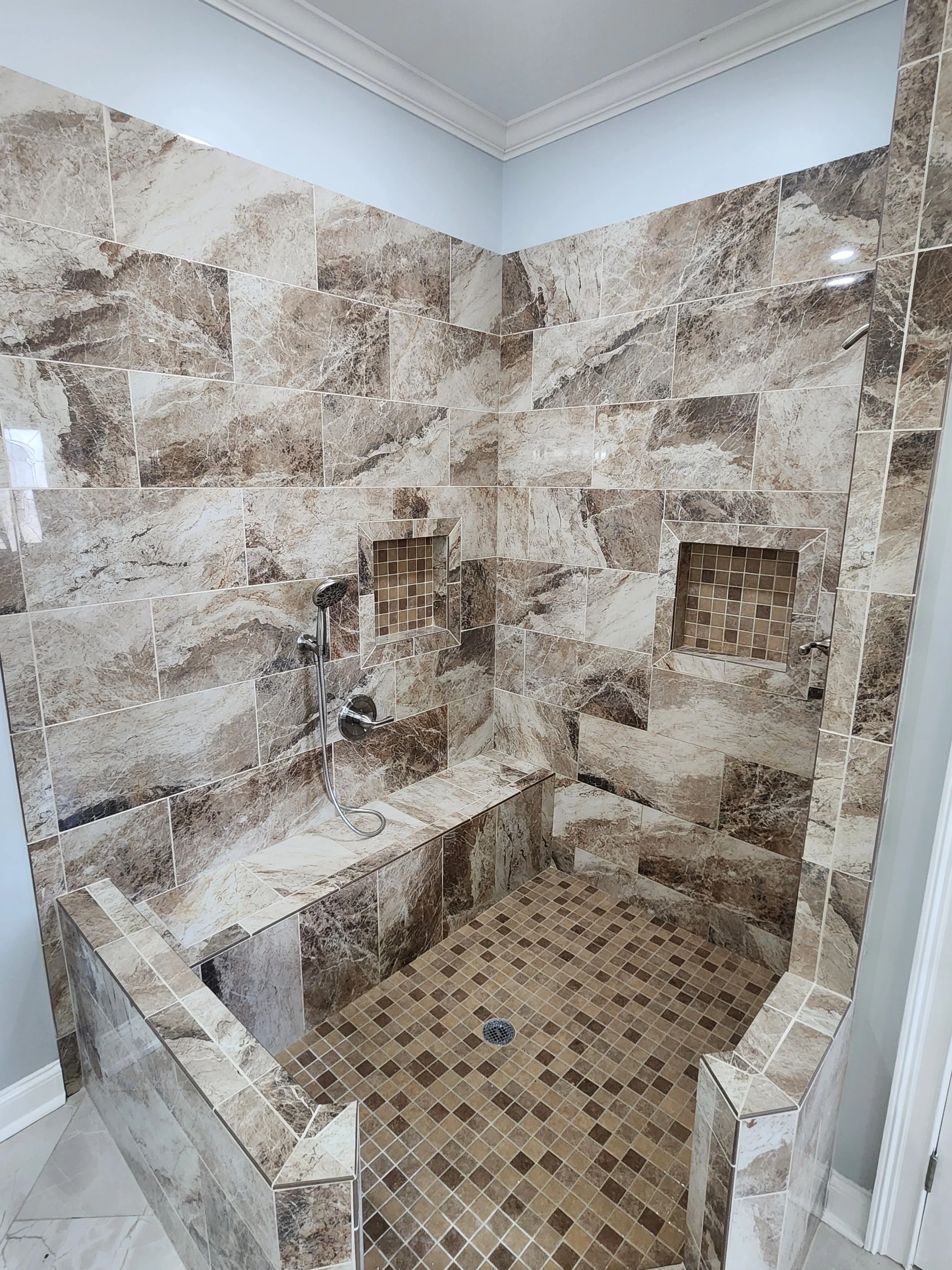 A walk-in shower with beige and brown marble tiles, built-in tiled niches, and a small bench. The bathroom has light blue walls and white crown molding.