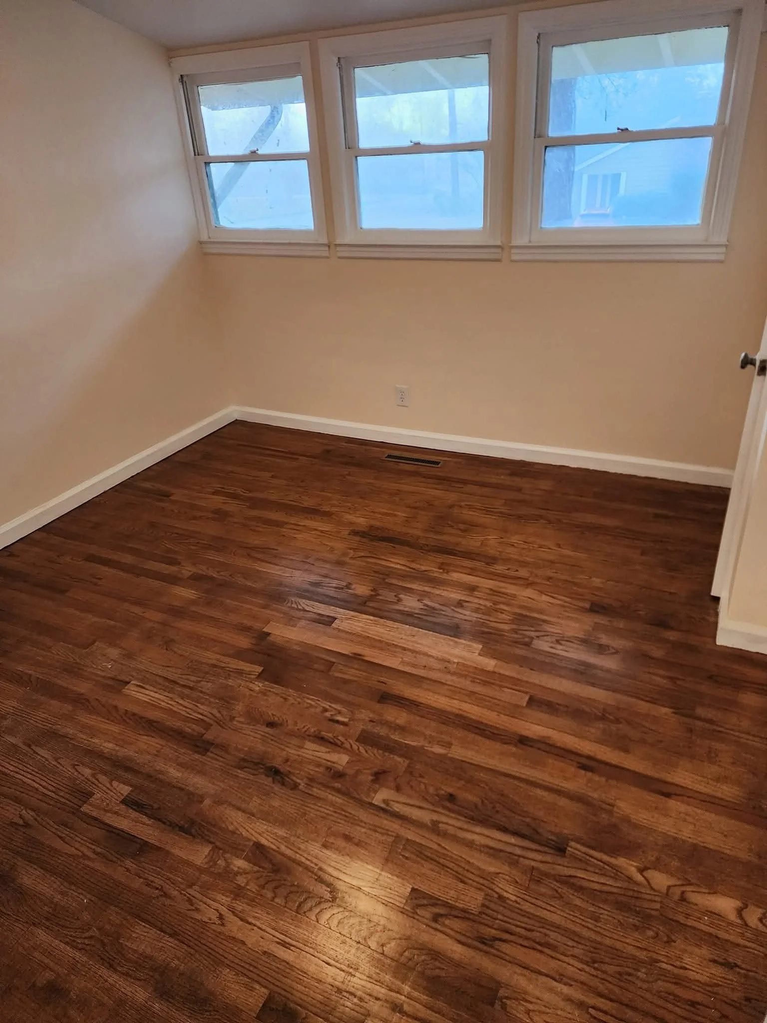 Empty room with hardwood floors, beige walls, and four windows with white frames that look outside to a tree and neighboring house.