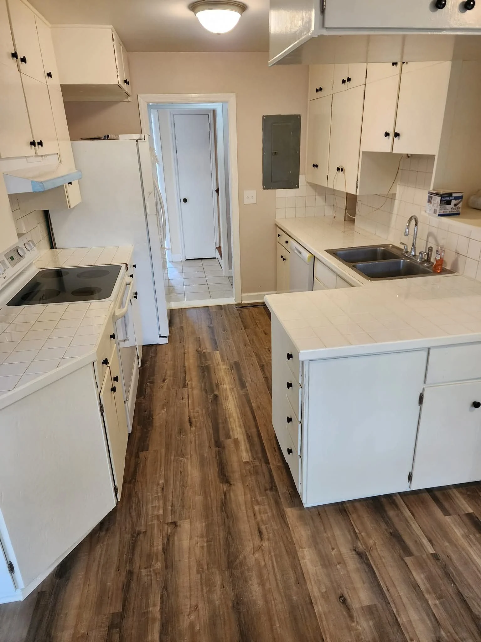 Empty kitchen with white cabinets, wood floor, double sink, electric stove, and fridge, with a doorway leading into a small tiled room.