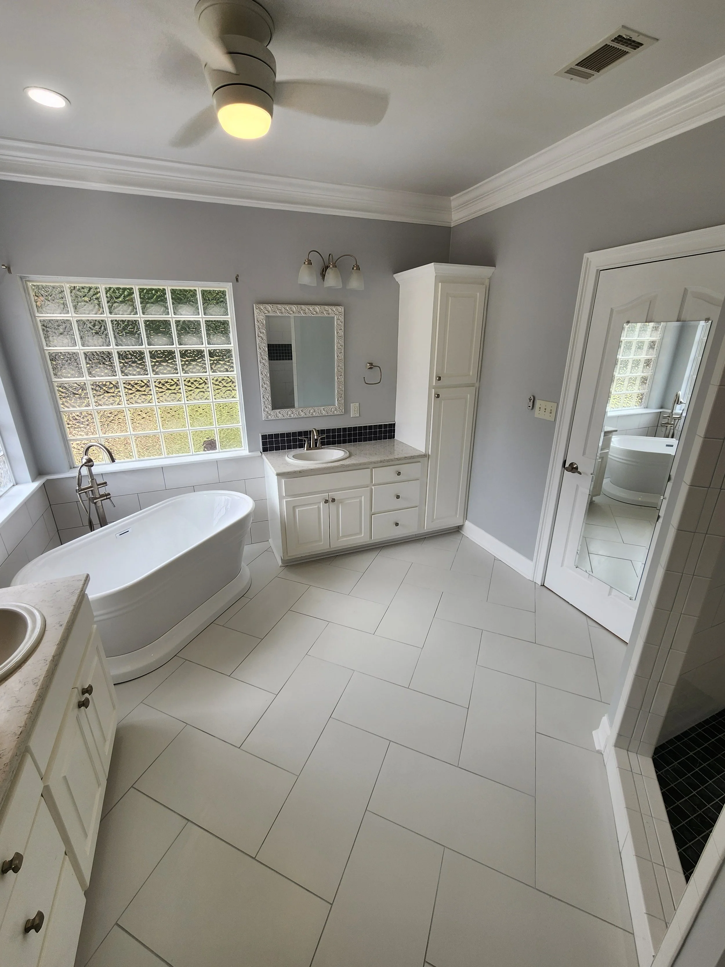 A bright bathroom with white tiling, a freestanding bathtub by a large glass block window, white cabinets, and a mirror above a small sink. There is a ceiling fan and a door with a mirror leading to a shower area.