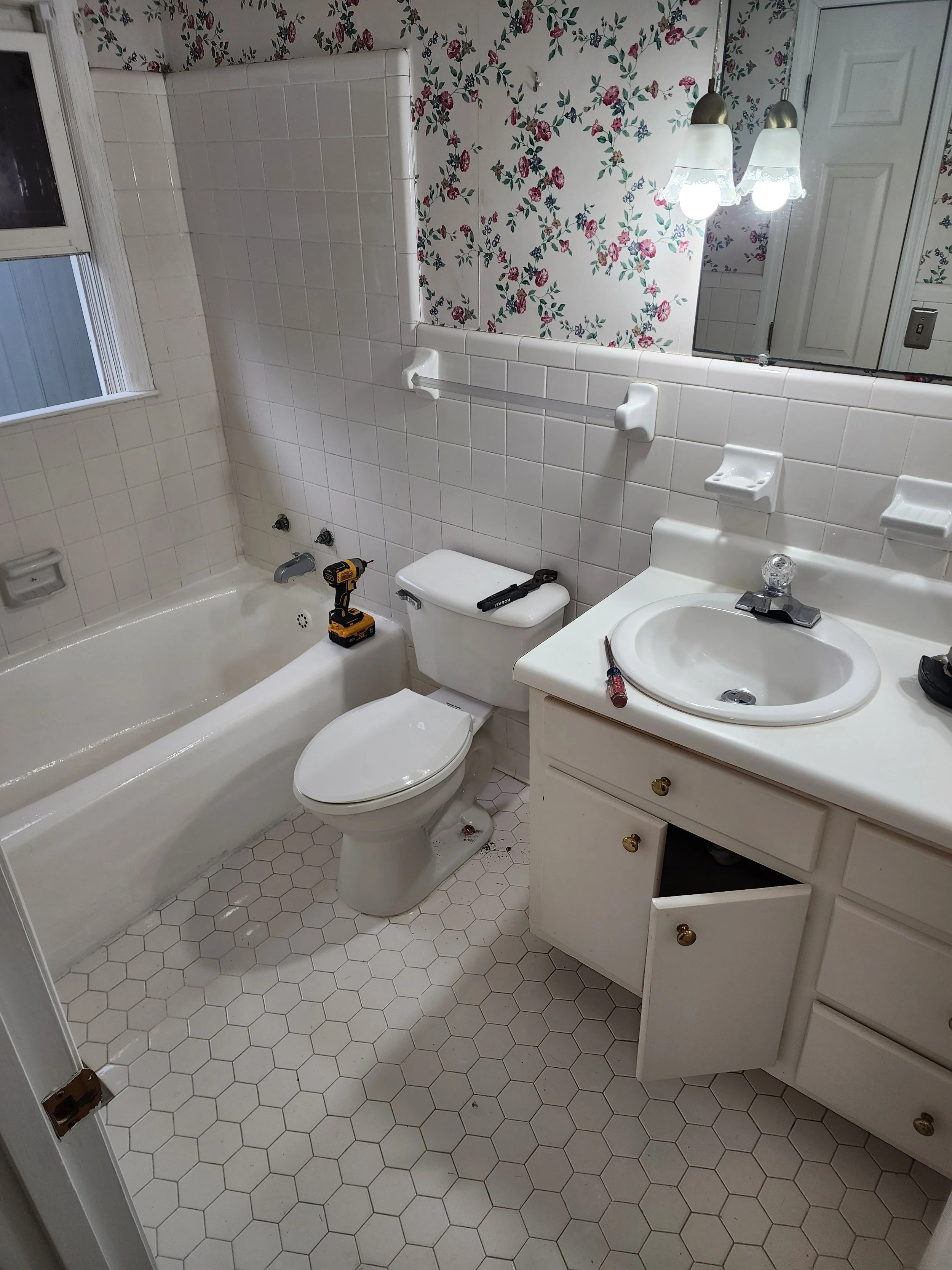 A small bathroom with a bathtub, toilet, sink, and mirror. The wall has floral wallpaper, and there are tools on the toilet and sink, indicating ongoing repairs.