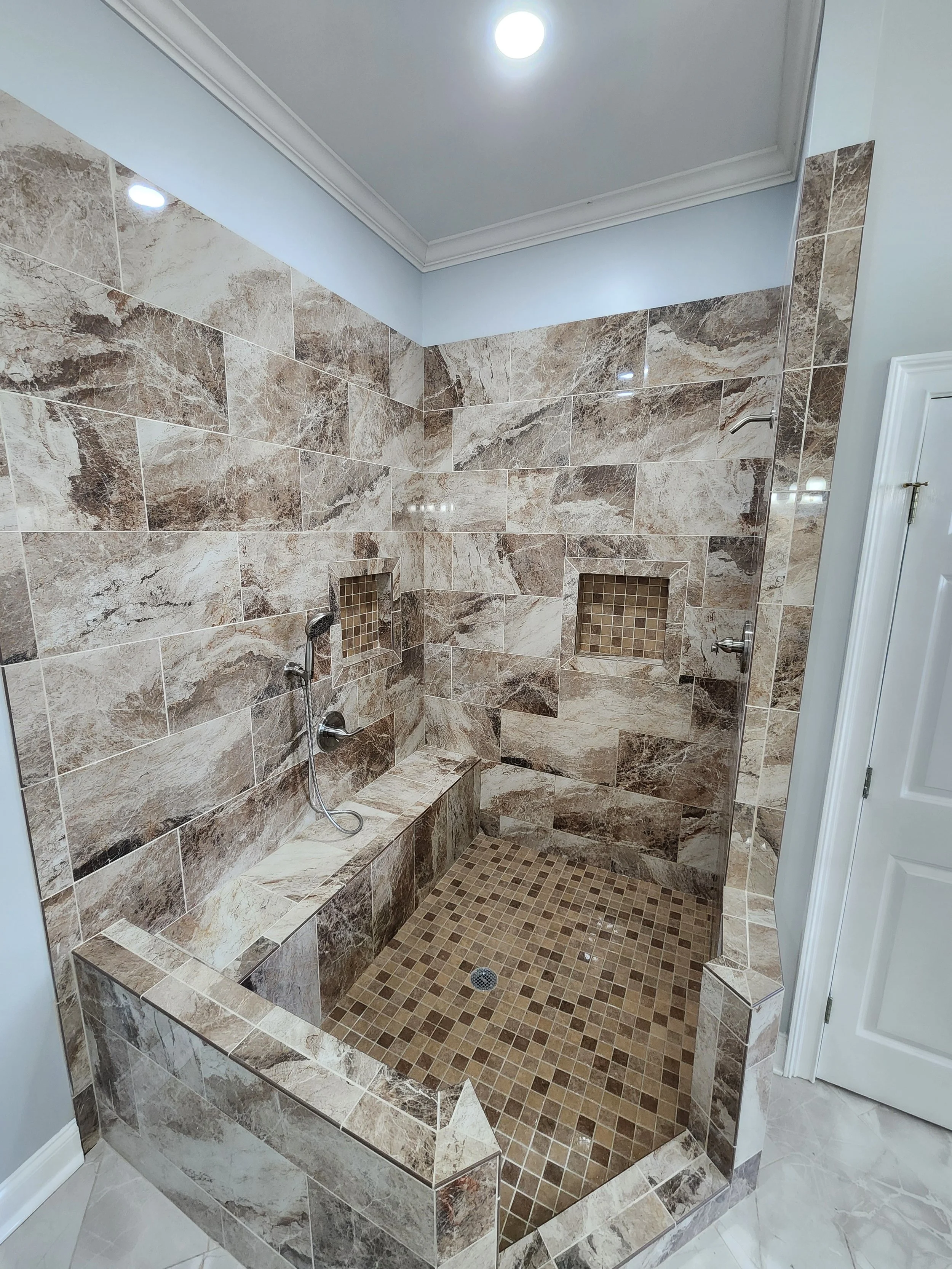 A walk-in shower with brown and beige marble tiles, a built-in marble bench, dual wall niches with small mosaic tiles, a handheld showerhead, and a ceiling light.