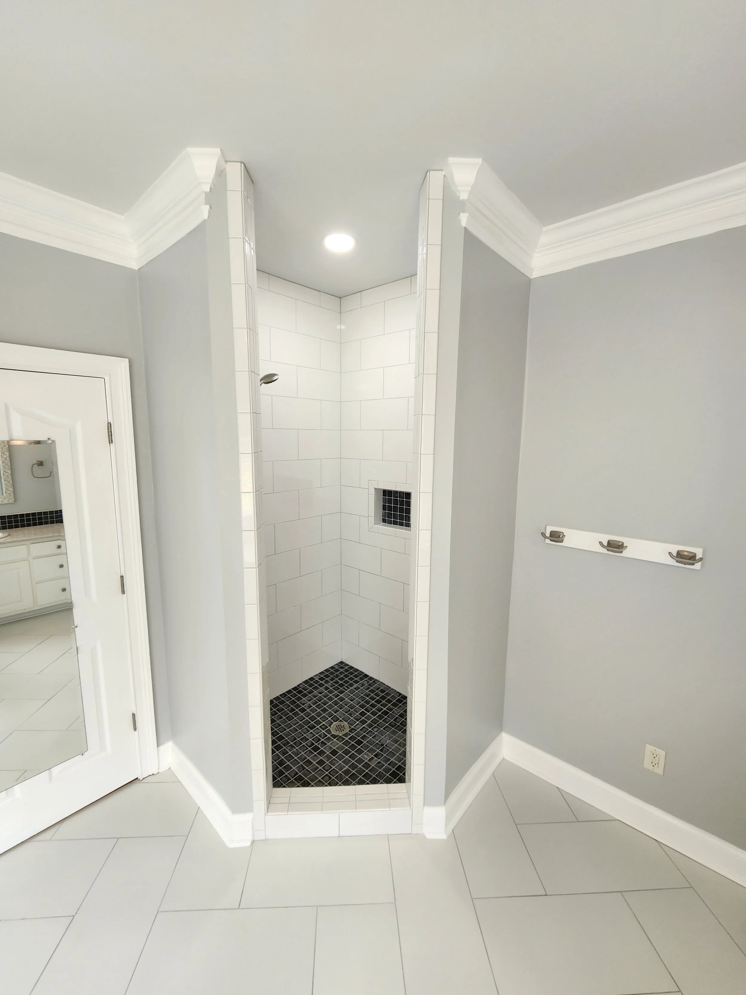 Small walk-in shower with white tiles and black accent tiles on the floor and a small black tiled niche. The shower is in a corner with grey walls and white crown molding.